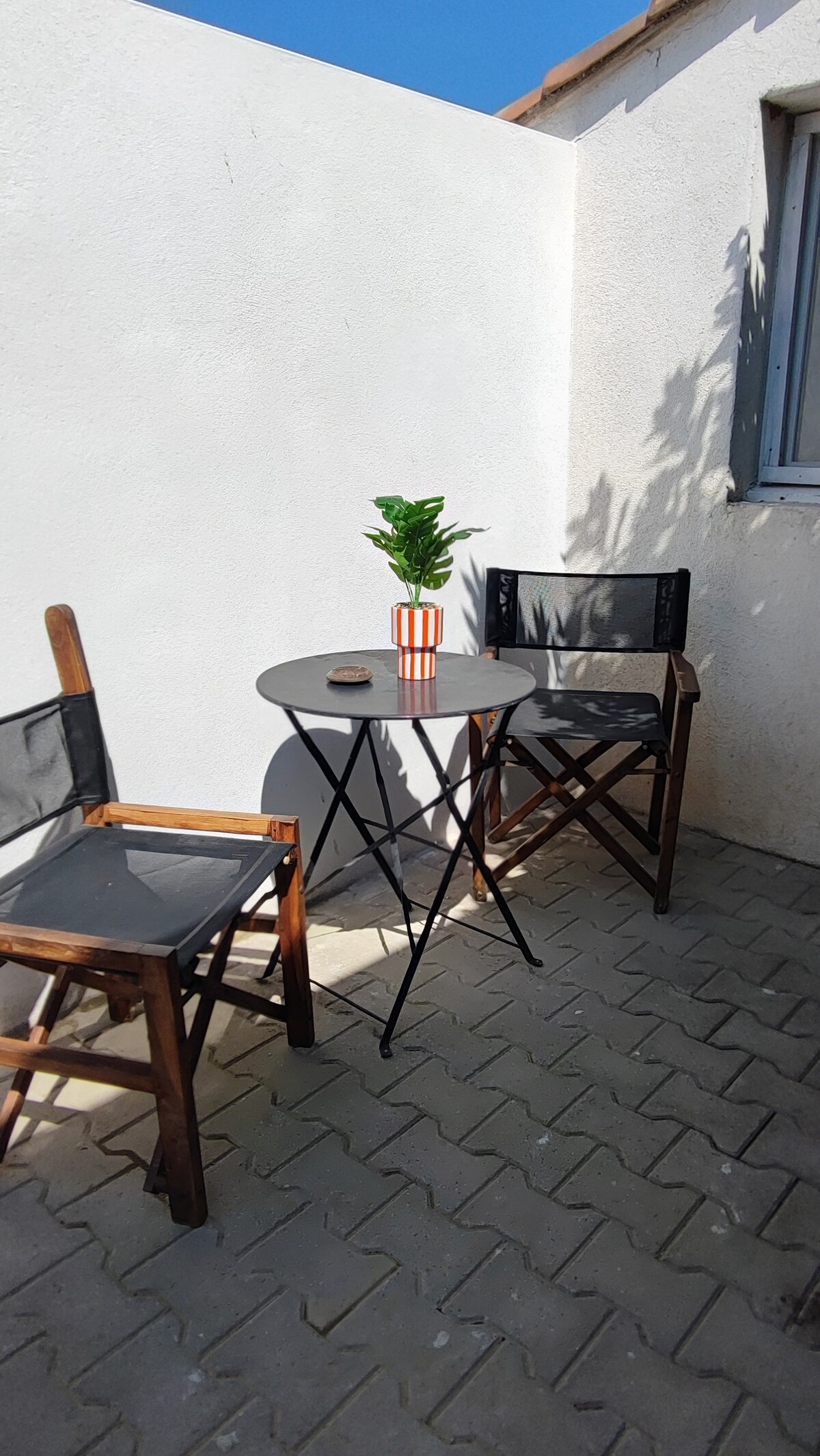 An outdoor seating area is presented with two wooden chairs and a small round table. A plant in a striped pot adds a decorative touch, while sunlight casts gentle shadows on the wall. The setting offers a private spot for relaxation.