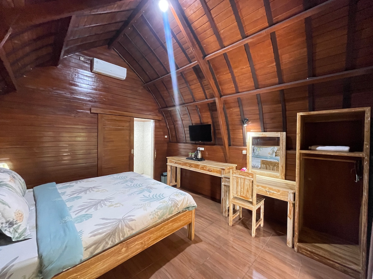 A wooden-beamed room features a spacious bed adorned with light-colored bedding. A wooden desk and chair occupy one side, accompanied by a small television mounted on the wall. A closet provides storage, and soft lighting enhances the warm atmosphere.