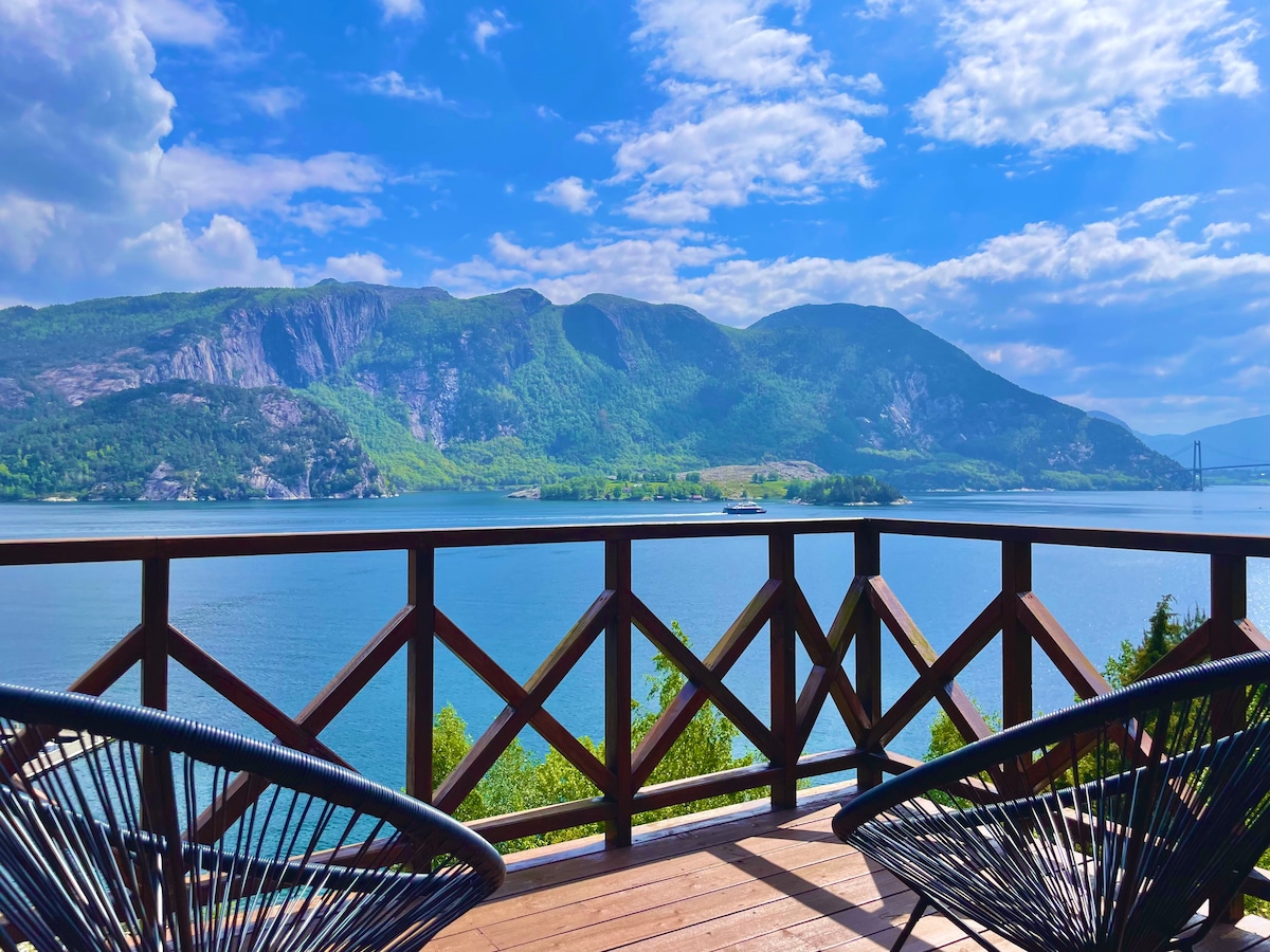 A wooden balcony overlooks a serene fjord, with lush green hills rising in the background. Two stylish chairs provide a comfortable seating option. The water reflects the sky's blue hues, while clouds drift gently above, creating a peaceful setting.