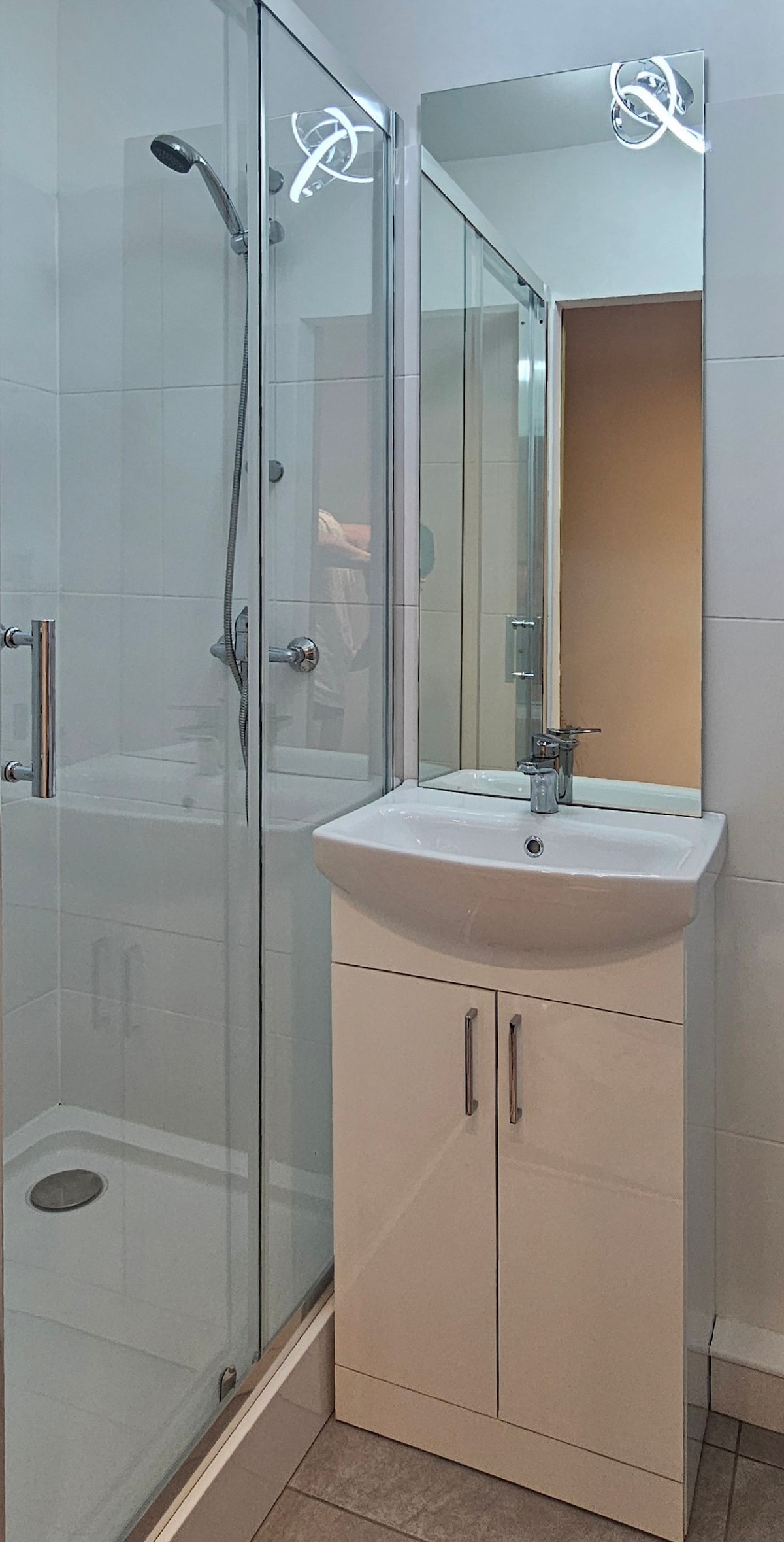 A modern bathroom featuring a glass-enclosed shower with a sleek chrome showerhead. A compact vanity with a white sink and storage cabinets below is positioned next to a large mirror, enhancing the sense of space and light.