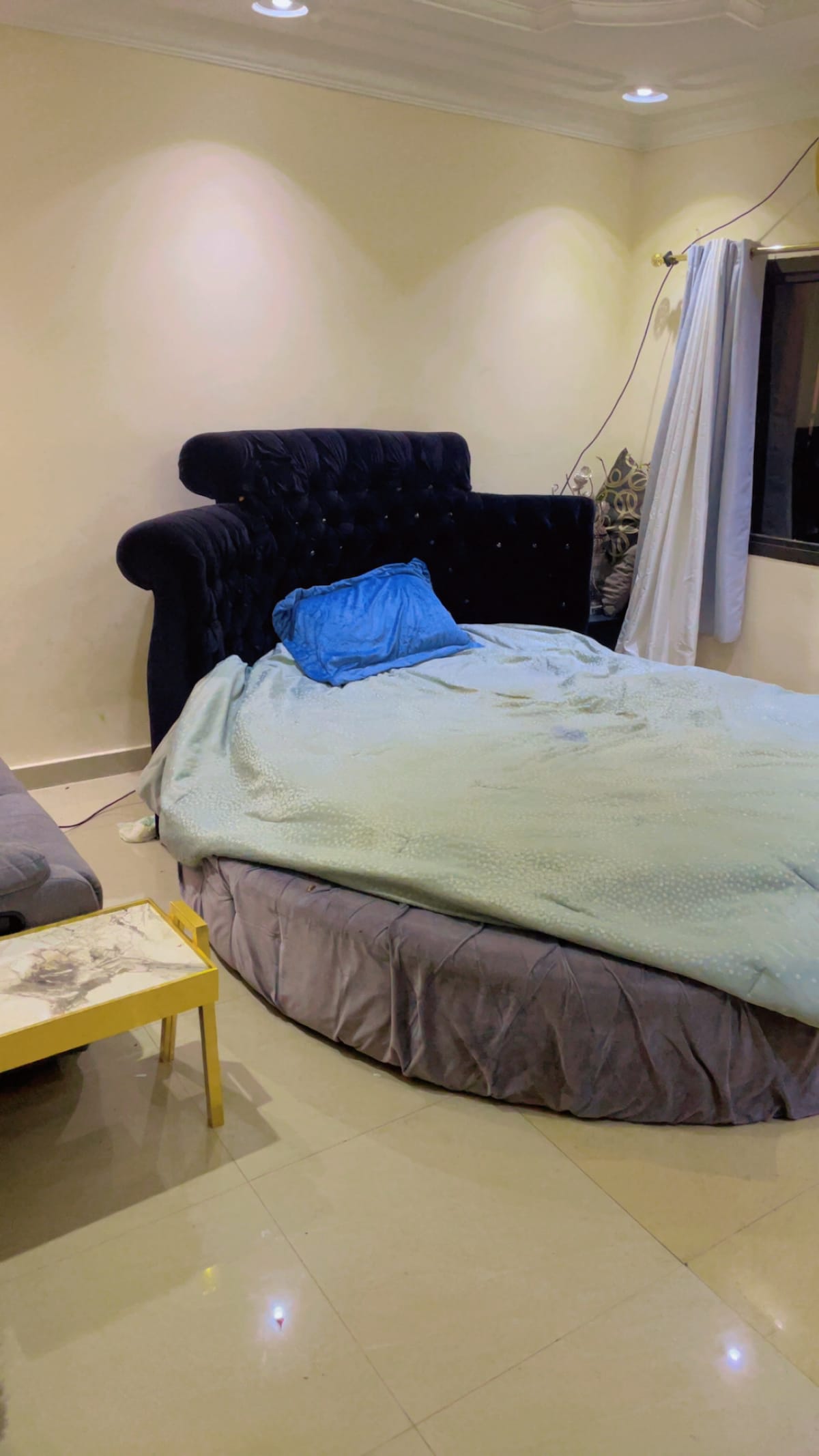 A spacious sleeping area features a large, round bed with a light blue blanket draped over it. A dark velvet headboard provides a plush backdrop. Nearby, a gray sofa offers additional seating, next to a small, light-colored table. Natural light enters through a window covered by sheer curtains.