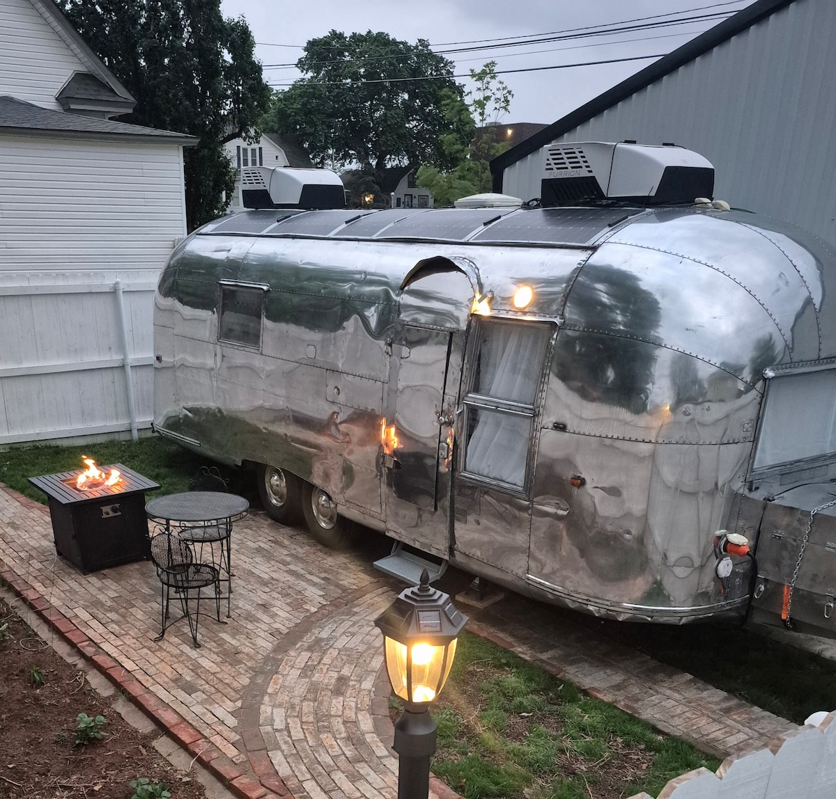 The vintage Airstream exterior is displayed, reflecting its polished metal surface. A seating area with two chairs and a small table is visible near a fire pit. Soft outdoor lighting from a nearby lamp creates a warm ambiance in the fenced backyard.