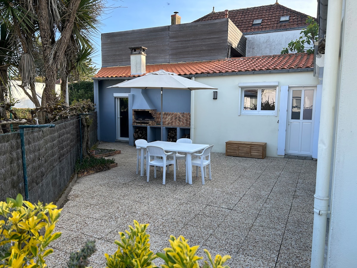 An outdoor courtyard features a dining table surrounded by white chairs, with a large umbrella providing shade. A barbecue area is positioned adjacent to the table. Lush greenery is visible along the perimeter, enhancing the outdoor living space.