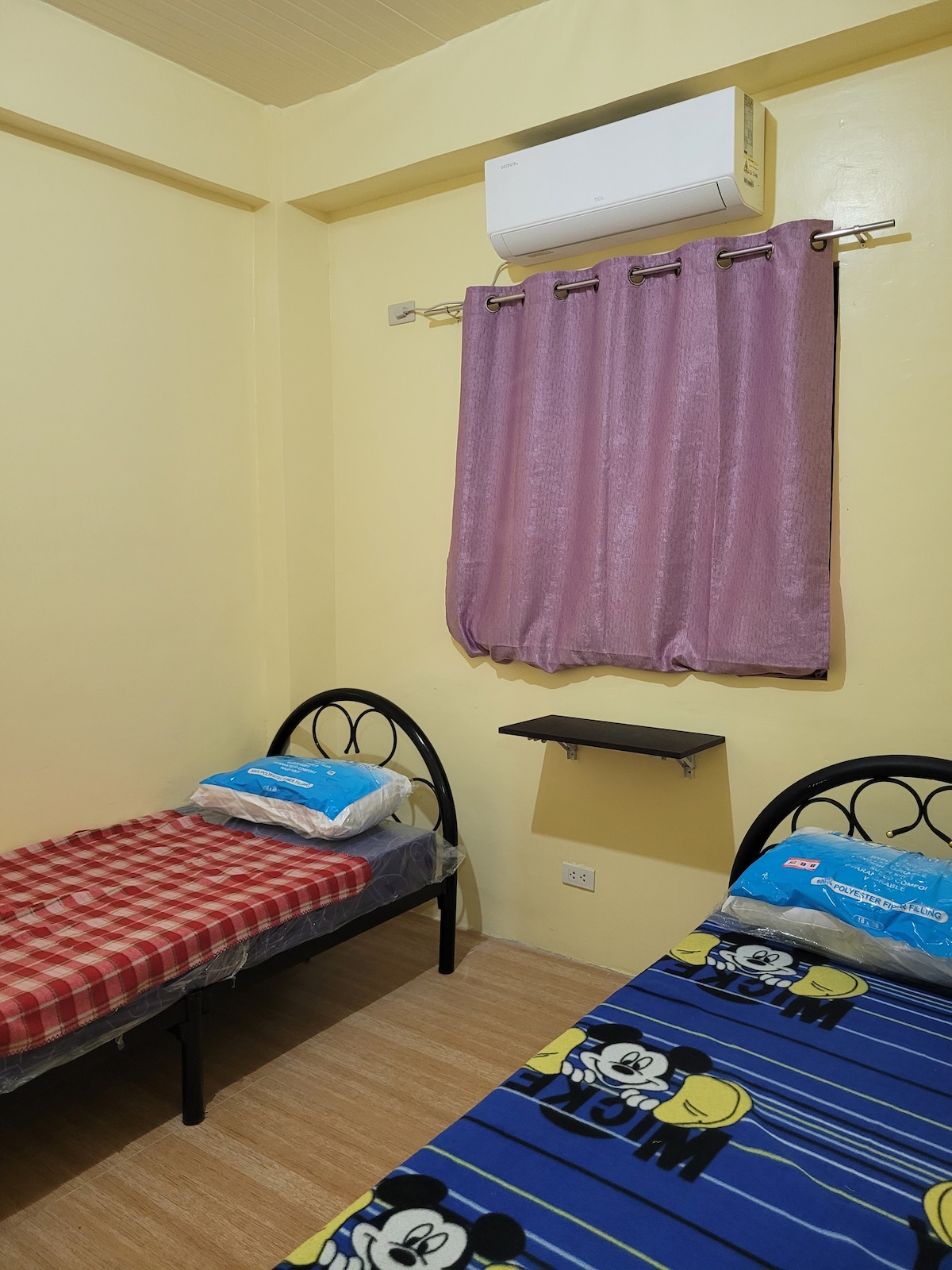 A bedroom is depicted with two single beds, one draped in a red-and-white plaid bedding and the other in a blue Mickey Mouse-themed cover. Light-colored walls and a window covered by a sheer purple curtain are visible. A small shelf is mounted on the wall between the beds.