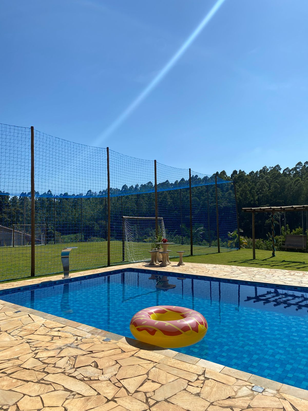 A swimming pool is surrounded by a fenced patio, featuring a colorful float resting on the water's surface. Sunlight reflects off the clear water, with green hills visible in the background. The stone-tiled area provides ample space for relaxation.