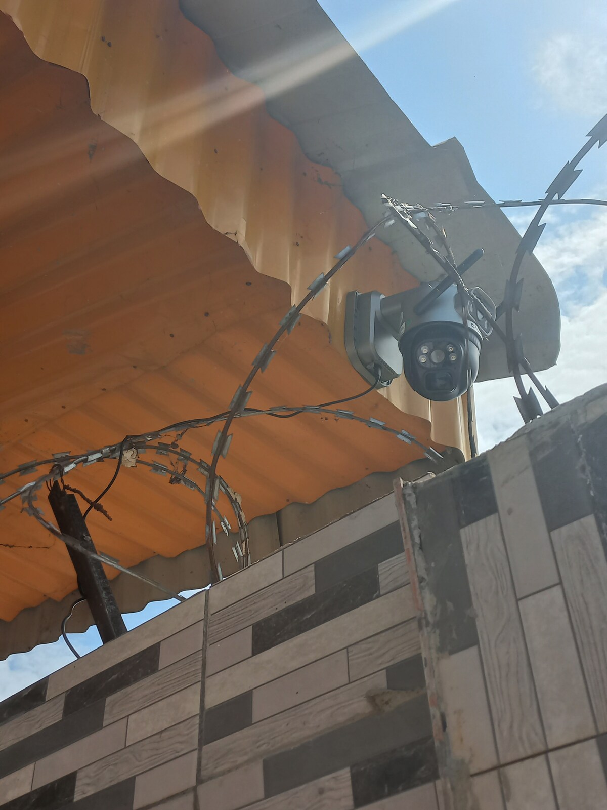 A security camera is mounted on a wall under an orange corrugated roof. Barbed wire is visible along the top edge of the boundary wall, which features a design of alternating light and dark tiles. Sunlight creates a bright atmosphere in the image.