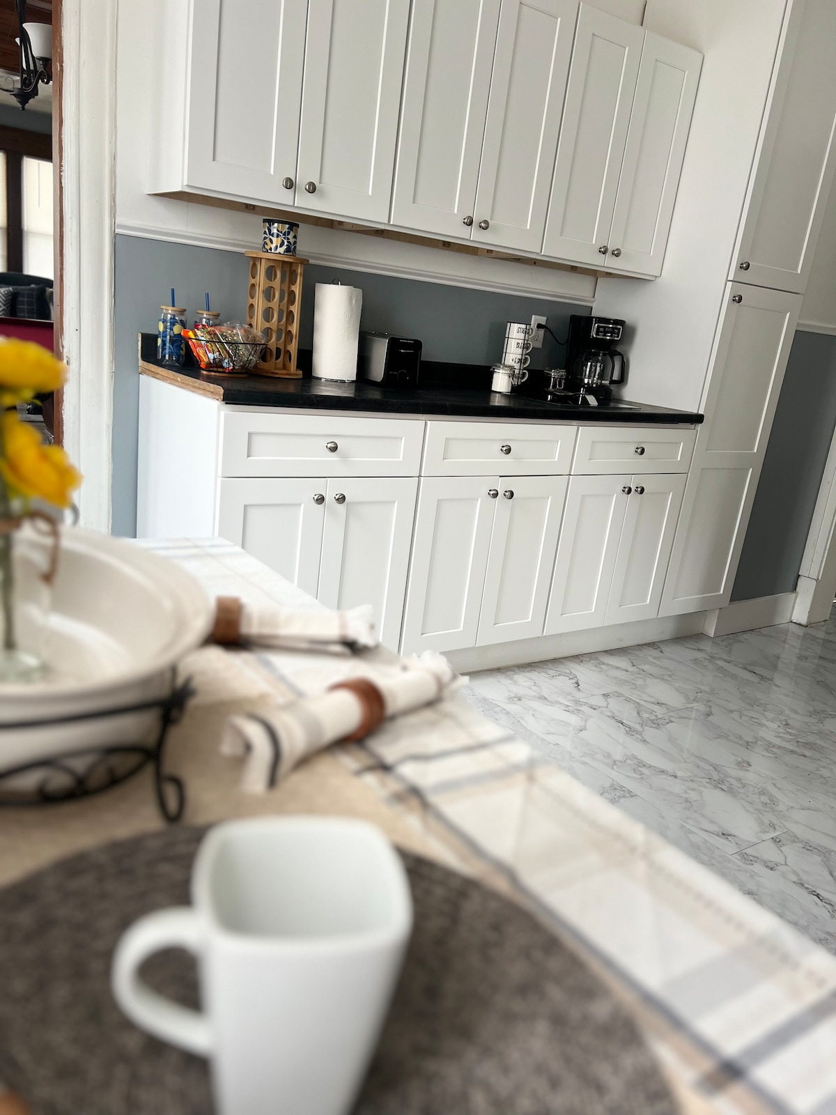 A clean and modern kitchen is showcased, featuring white cabinetry and a dark countertop. A coffee maker and a sleek coffee machine are visible, alongside storage for utensils. The space is complemented by a marble-like floor and a dining area set with a placemat and cup.