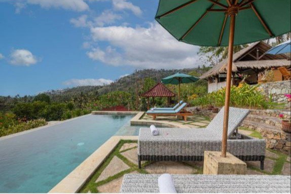 A serene pool area is surrounded by greenery, featuring two comfortable loungers under large green umbrellas. The infinity pool extends towards scenic views of the hillside, with stone pathways and well-kept landscaping enhancing the tranquil setting.