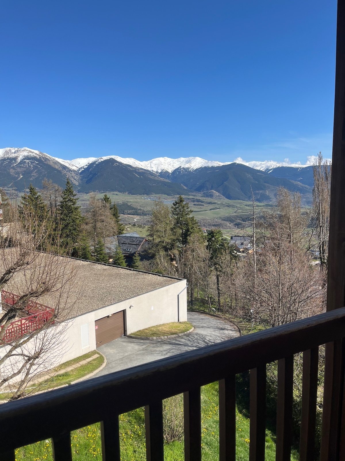 A scenic view is captured from a balcony, overlooking lush green hills and majestic snow-capped mountains under a clear blue sky. Nearby trees and the silhouette of a building can be seen, offering a sense of tranquility and connection to nature.