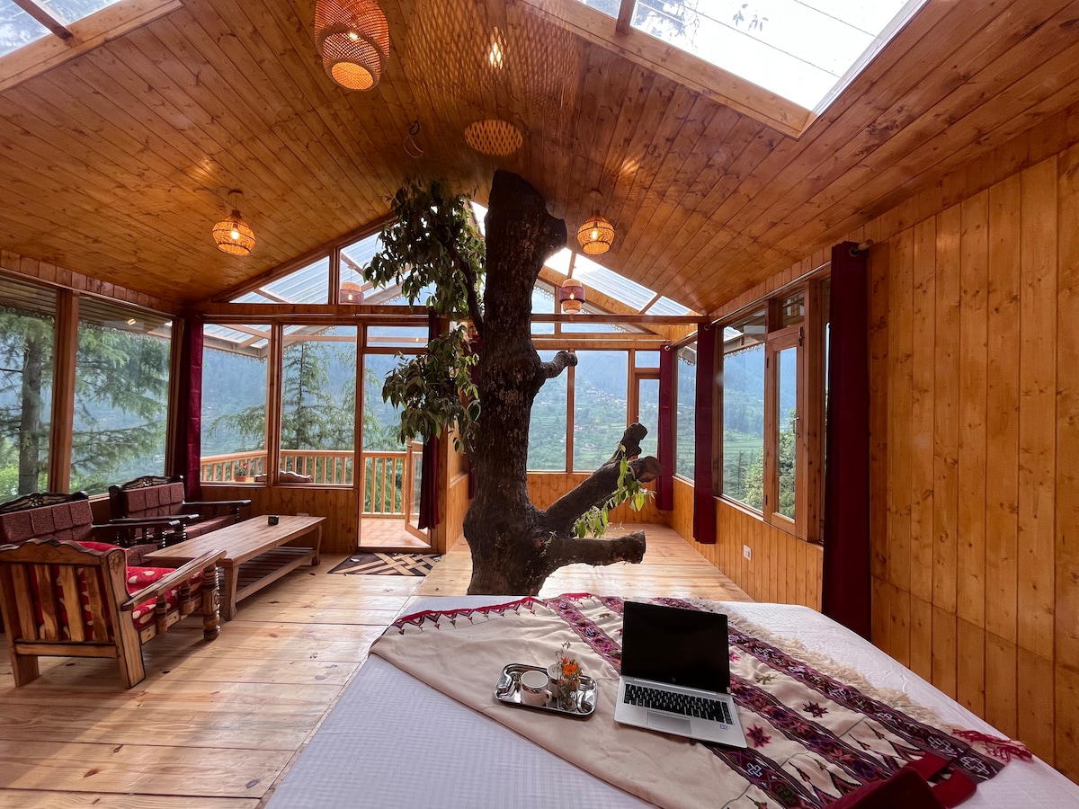An expansive treehouse interior features an oak tree integrated into the design, creating a unique focal point. Large glass windows offer views of the surrounding valley and invite natural light. A cozy bed is dressed with decorative throws, while wooden furnishings complement the warm wood paneling.