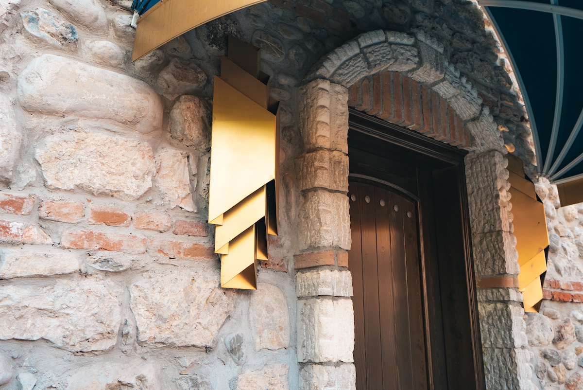 An entranceway is framed by a rustic stone wall, featuring warm-toned bricks and textured surfaces. A distinctive golden fixture is mounted beside the door, showcasing a geometric design that enhances the architecture. The wooden door is adorned with decorative paneling and subtle details.