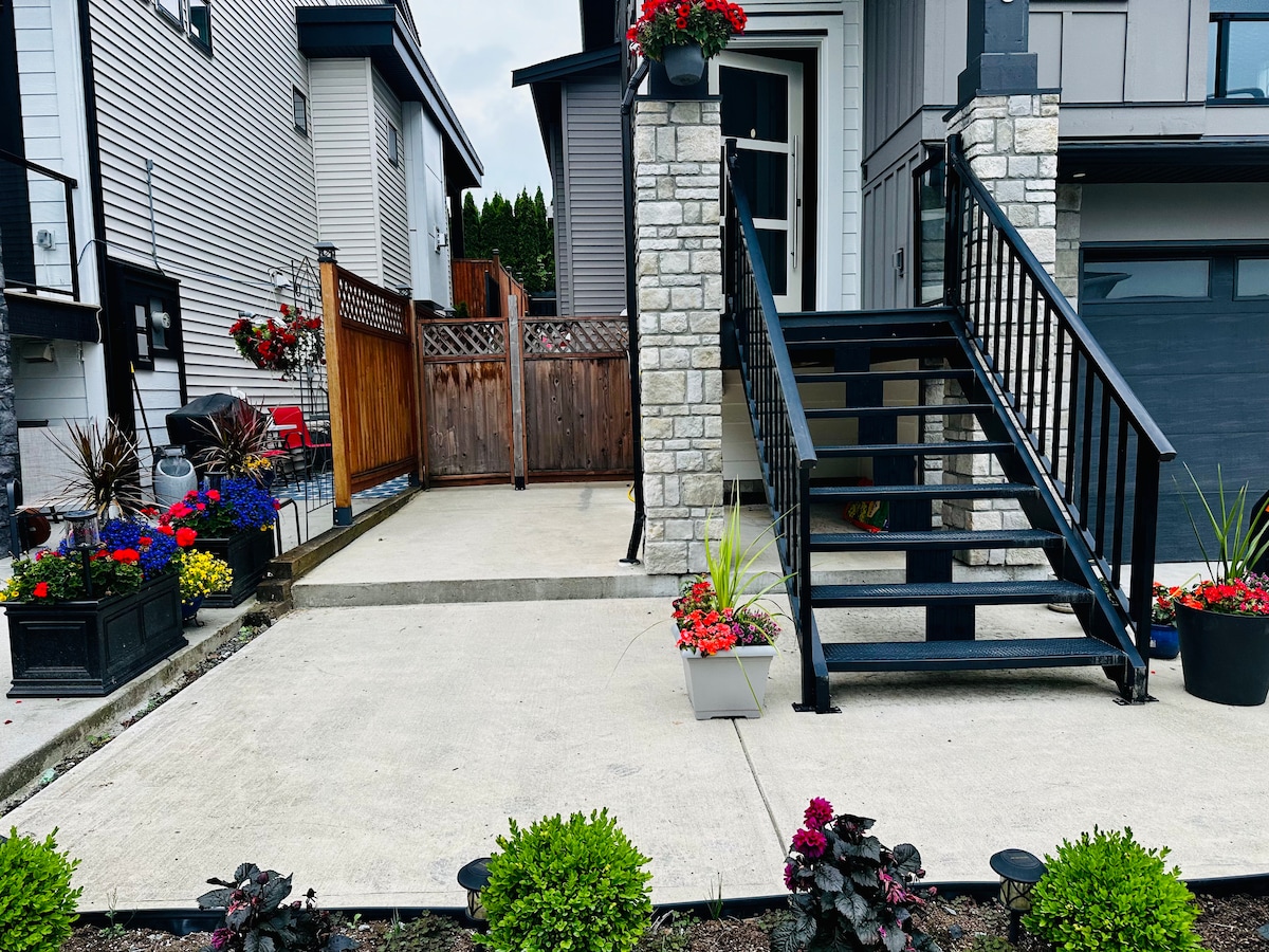 The exterior entrance showcases a well-maintained patio area with potted plants and vibrant flowers in various colors. A set of stairs leads up to the home's entry, with a decorative stone column framing one side. The area is surrounded by a wooden fence and a clean sidewalk.