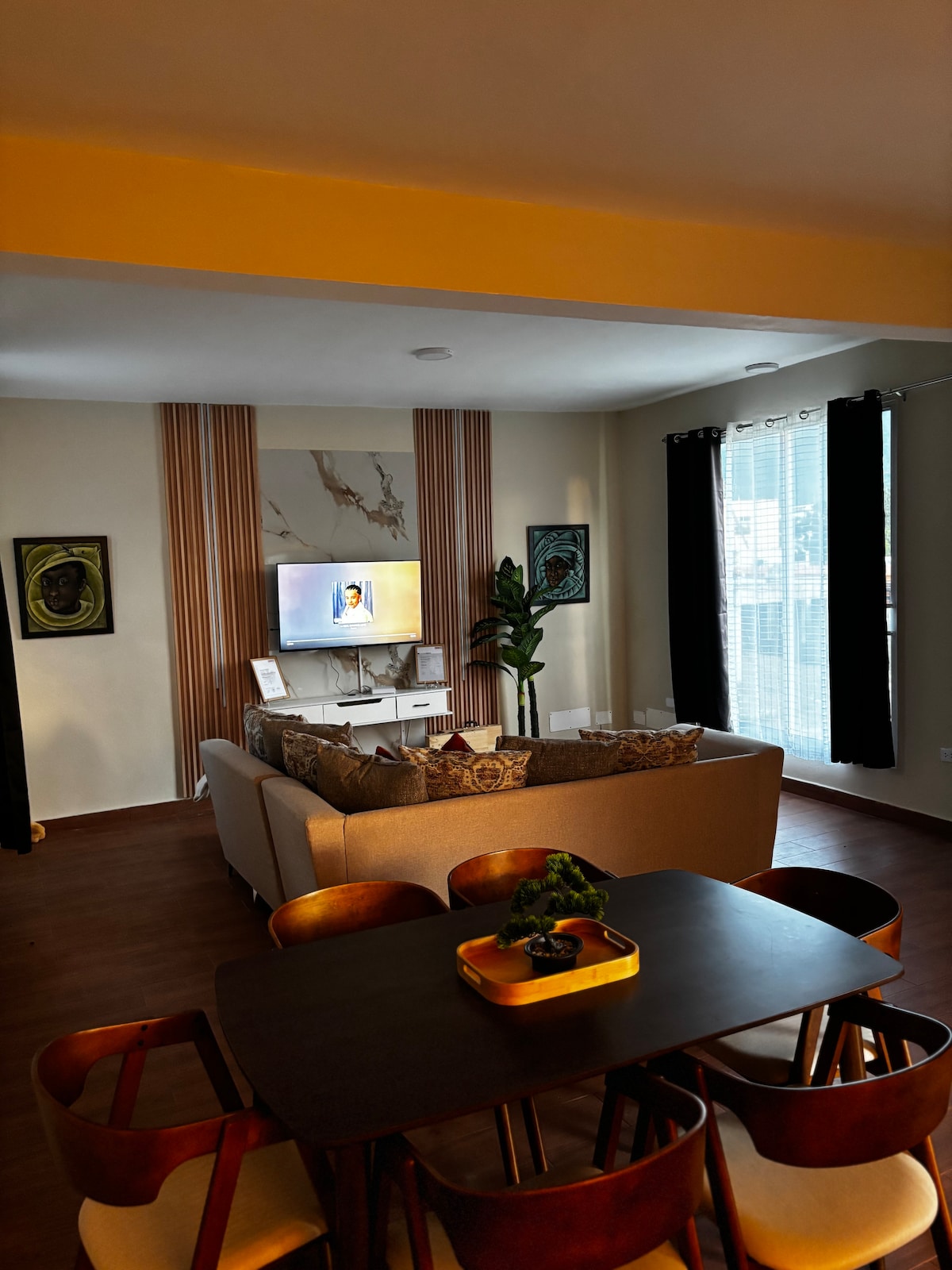 A cozy living area is displayed, featuring a spacious couch facing a wall-mounted television. Light curtains frame large windows, allowing natural light to fill the room. A dining table with wooden chairs occupies the nearby space, complemented by decorative wall art and indoor plants.