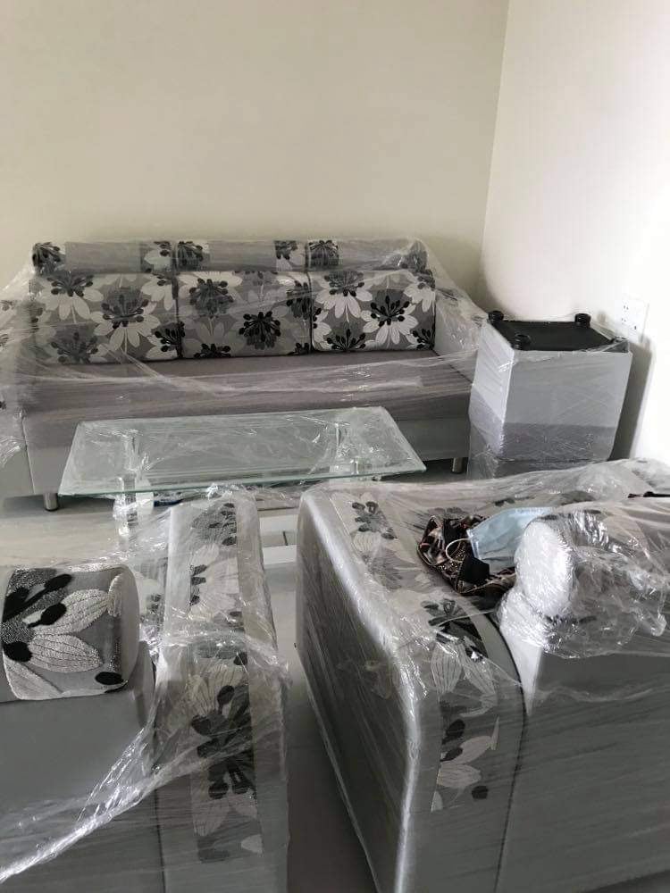 A modern living area is displayed, featuring a gray couch and armchairs with floral-patterned upholstery. A glass coffee table sits centrally, and the furniture is covered in protective plastic. Light-colored walls create a bright atmosphere around the seating arrangement.