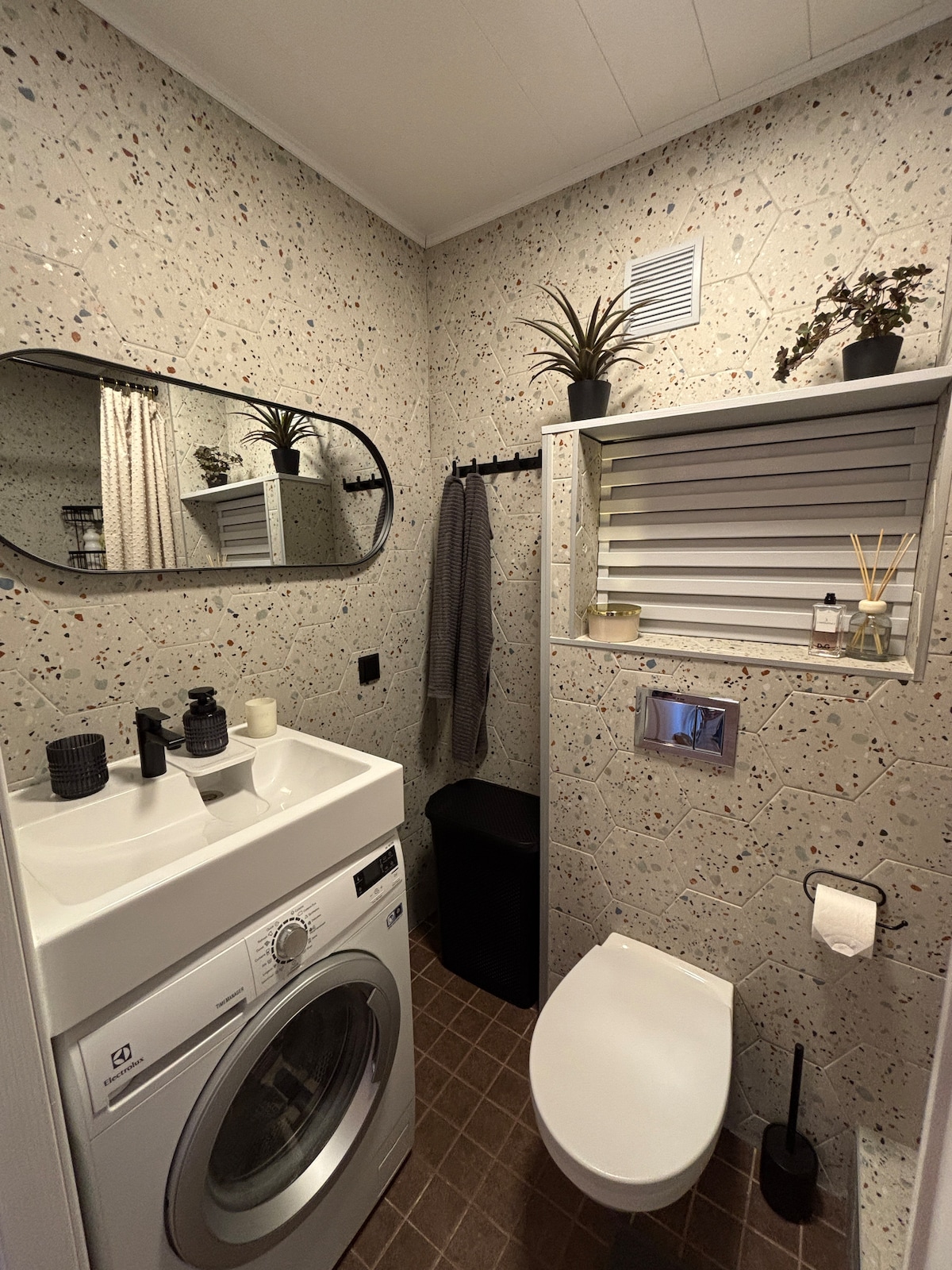 A modern bathroom features a washing machine integrated below a clean, white sink. Natural elements are introduced with potted plants on a shelf and a wall-mounted mirror. Textured, patterned walls provide visual interest, while accessories are neatly arranged on surfaces.