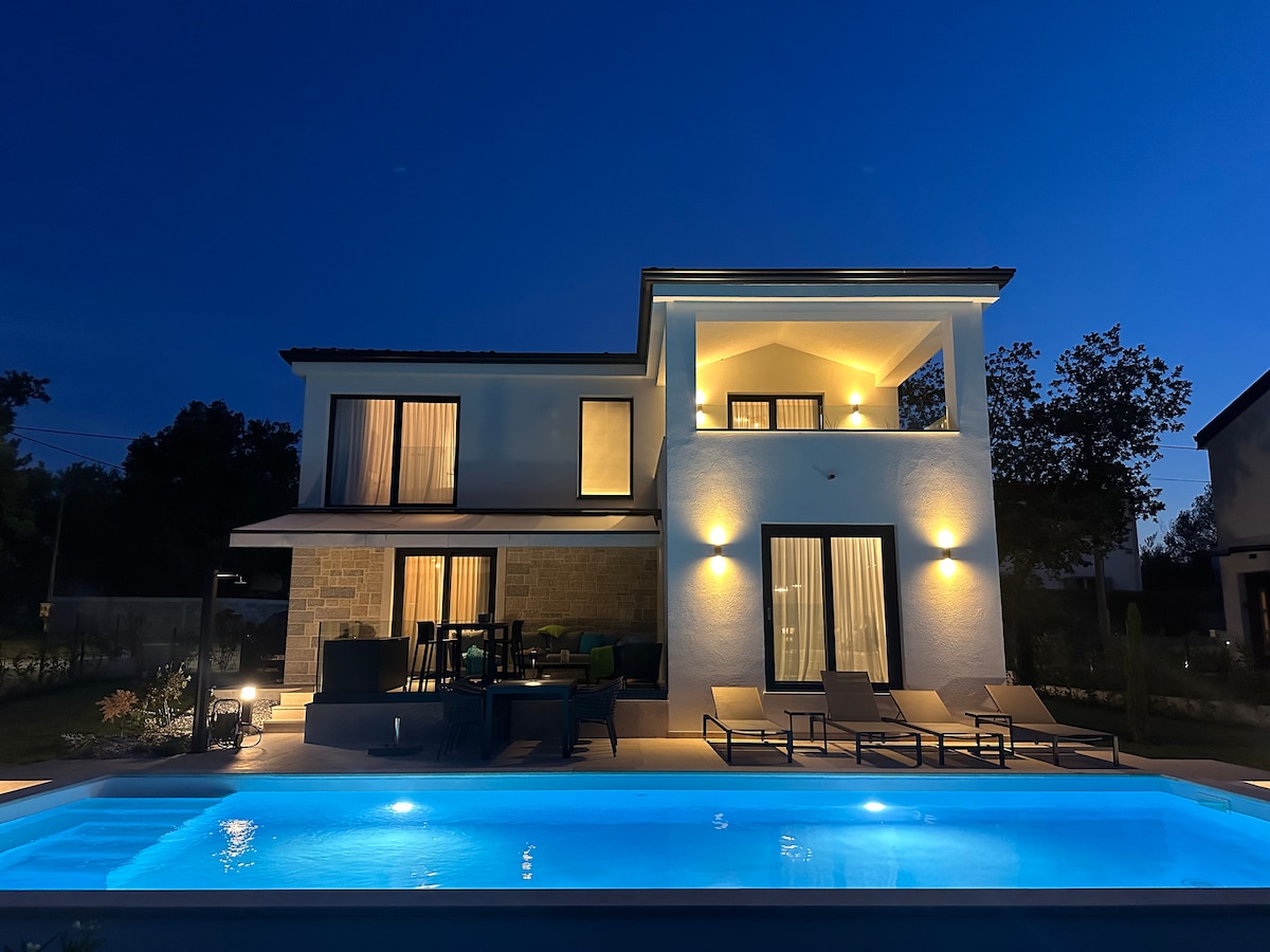 The villa is illuminated at night, highlighting its modern architecture and spacious terrace. A private pool reflects the tranquil blue water, surrounded by lounge chairs. Large windows allow for light to seep from the interior, enhancing the inviting atmosphere of the outdoor space.