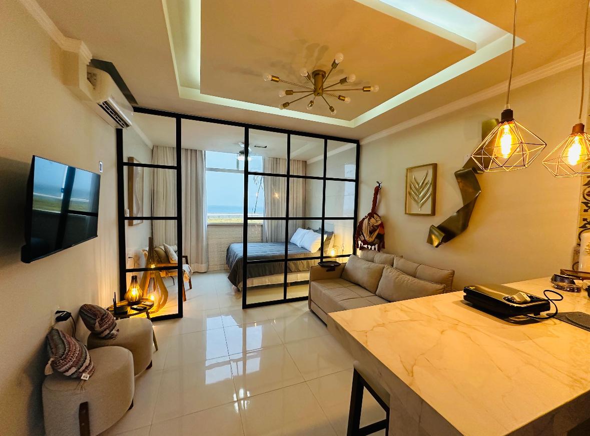 A studio apartment features a modern design with a glass partition separating the sleeping area from the living space. A spacious bed is visible behind the partition, while the living area includes a comfortable sofa and a dining table. Warm lighting is provided by pendant lights.