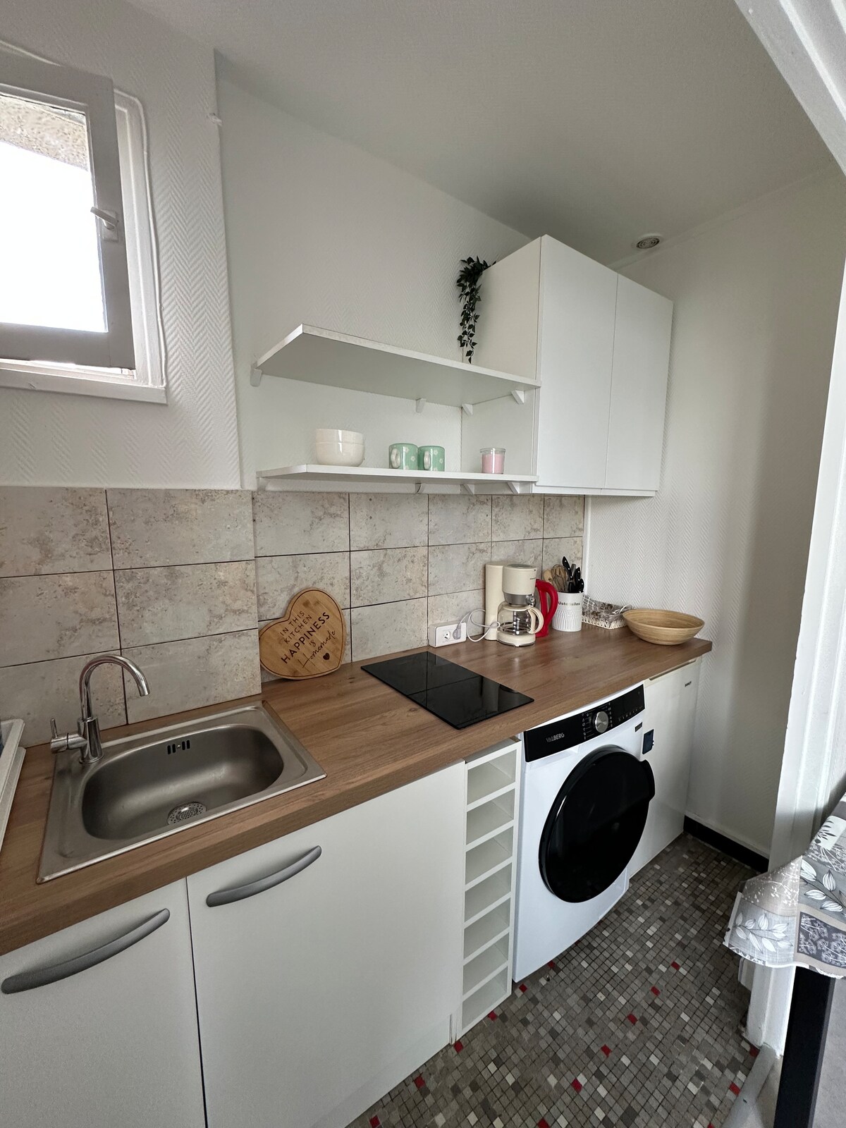 The compact kitchen features light cabinetry with open shelves and a wooden countertop. It includes a sink, an electric cooktop, and a washing machine. A window allows natural light to illuminate the space, while various kitchen items are neatly arranged on the shelves.