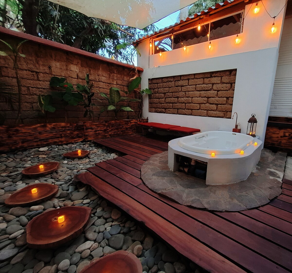 A serene outdoor space features a heated jacuzzi surrounded by smooth stones. Soft lighting from round candles adds a warm glow. Wooden decking frames the area, complemented by natural greenery and a stone wall in the background, creating a tranquil atmosphere for relaxation.
