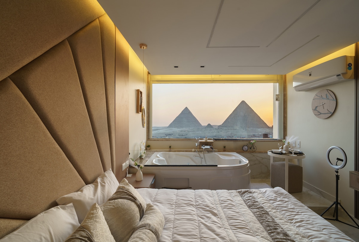A spacious bedroom is highlighted by a large, cushioned headboard. Natural light enters through a large window, revealing views of the iconic pyramids, while a modern bathtub is positioned nearby. Subtle lighting enhances the serene atmosphere, creating a relaxing space.