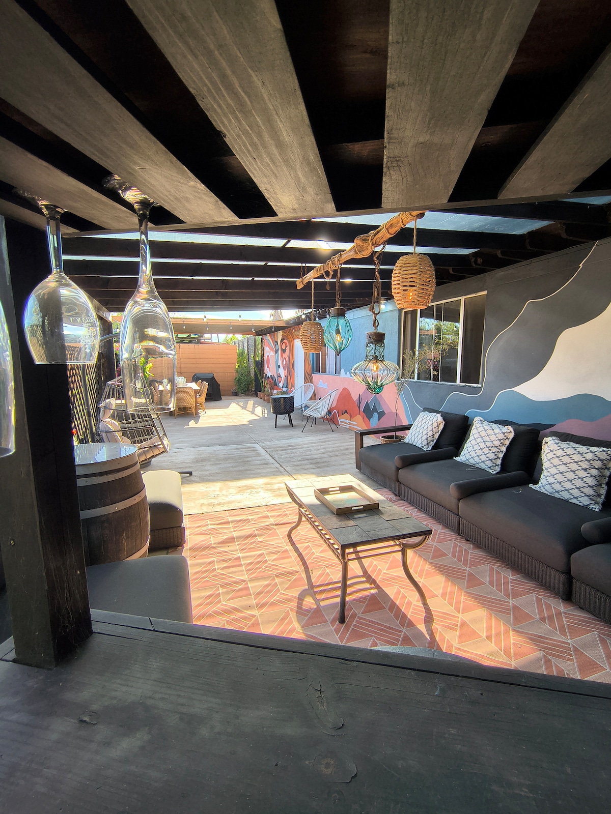 The colorful patio area features plush seating arranged around a coffee table. Hand-painted murals add visual interest to the surroundings, while several hanging glasses are visible above. A dining space is positioned in the background, designed for outdoor meals.