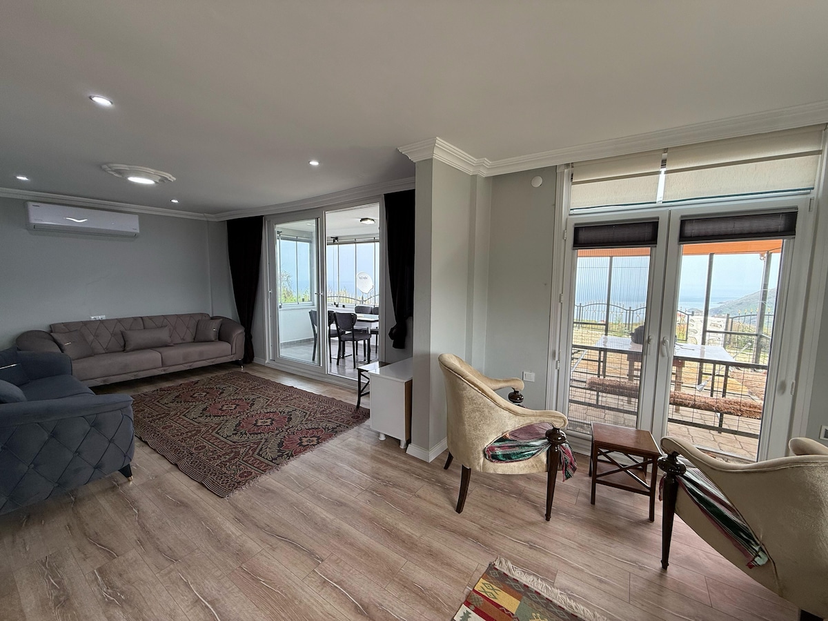 A bright living area is presented with modern furniture, including a gray sofa and two light-colored armchairs. Large glass doors offer views of the outdoor terrace, while a patterned area rug adds warmth to the wooden flooring. Air conditioning is visible on the wall.