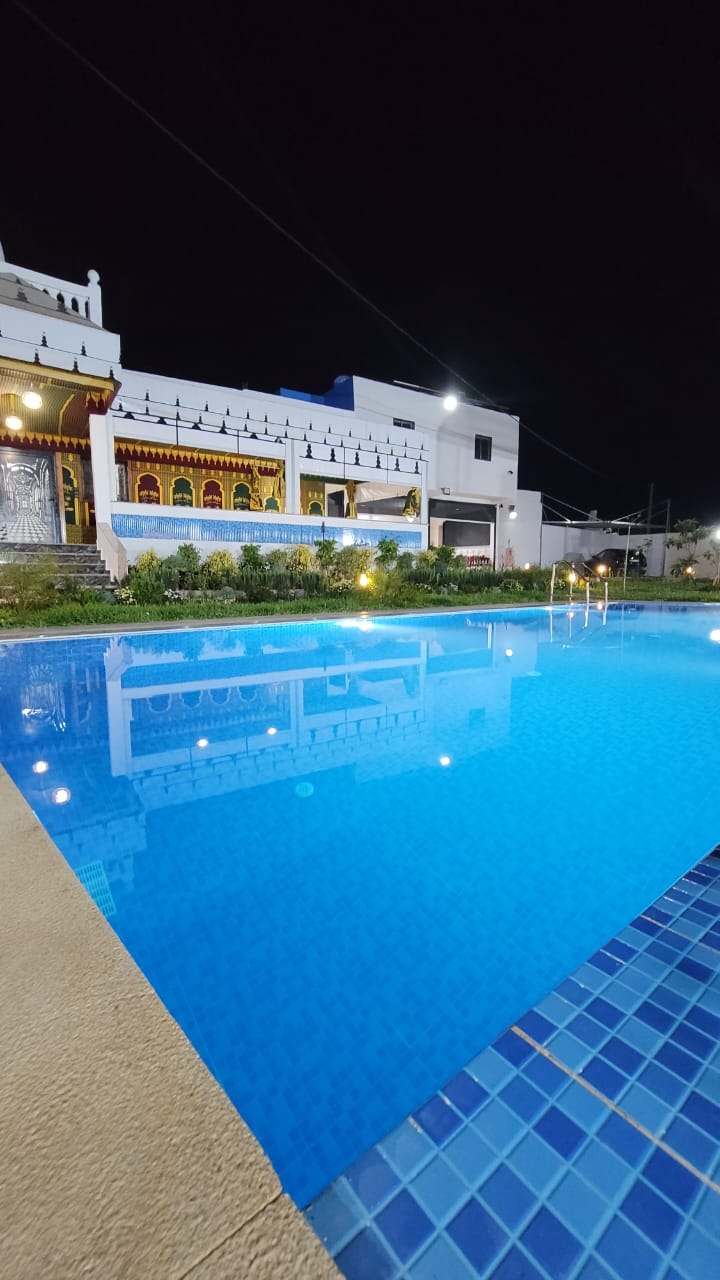 A private pool reflects the illuminated villa at night, surrounded by lush greenery. The water is a vibrant blue, and the villa features ornate architectural details and large windows that enhance the serene atmosphere.