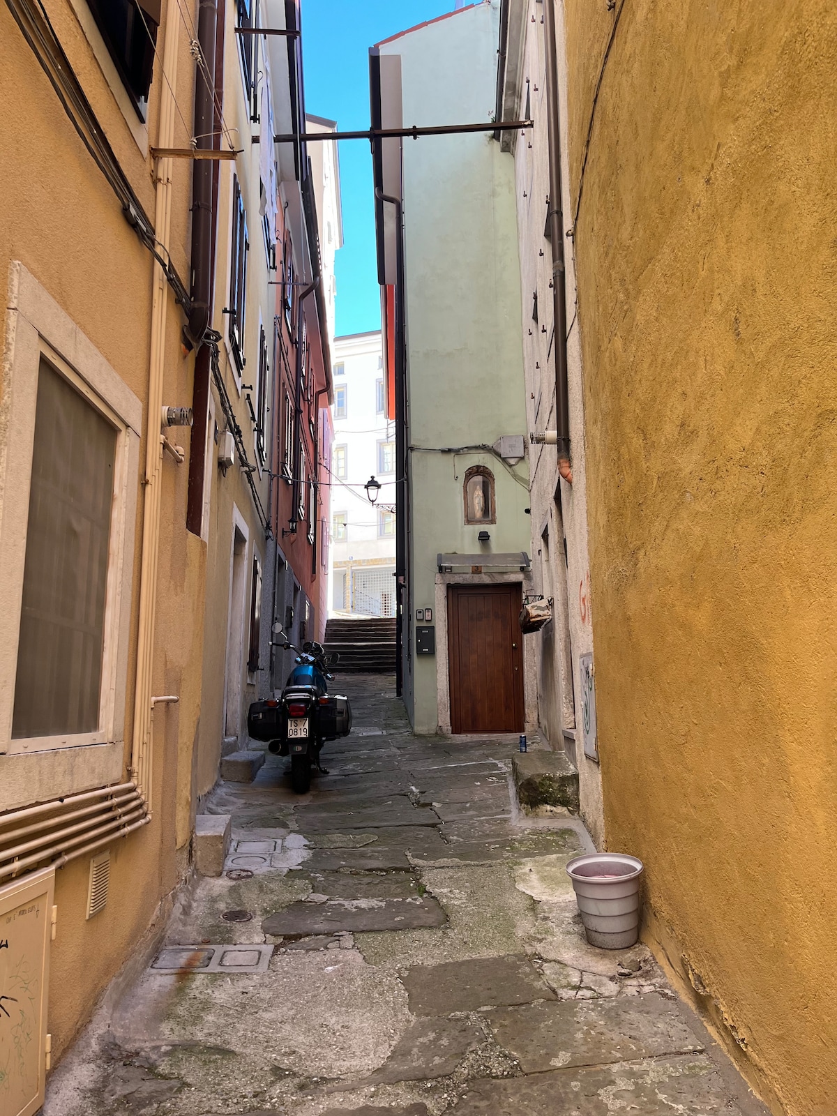 A narrow alleyway is framed by colorful buildings, with hues of yellow, green, and red visible on the walls. Stone cobblestones line the ground, leading towards a staircase in the background. A motorcycle is parked beside a white planter, adding to the urban atmosphere.