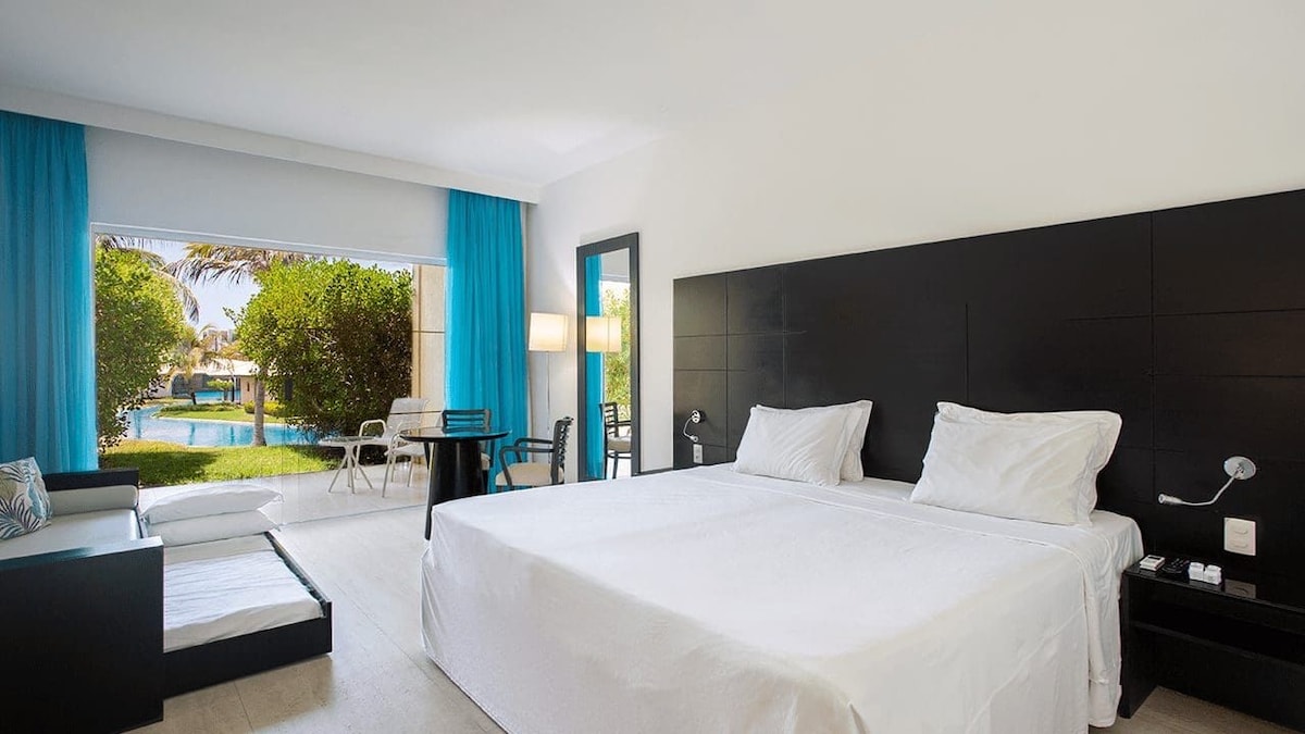 A spacious guest room features a large bed with white linens. Sliding glass doors lead to a lush outdoor area with greenery and a pool. A comfortable seating area with a small table is positioned beside the bed, enhancing the room's functionality.