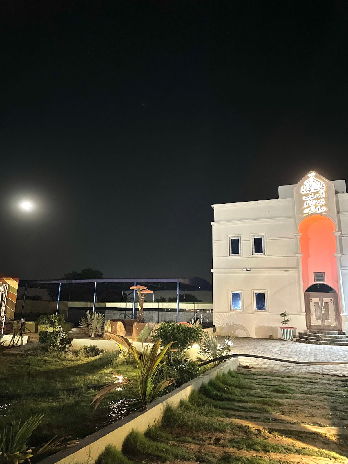 The exterior of the luxury farmhouse is illuminated at night, showcasing a white façade with decorative arches. A full moon is visible in the sky, adding to the serene ambiance. Landscaping features include plants and a gravel path leading toward the entrance.