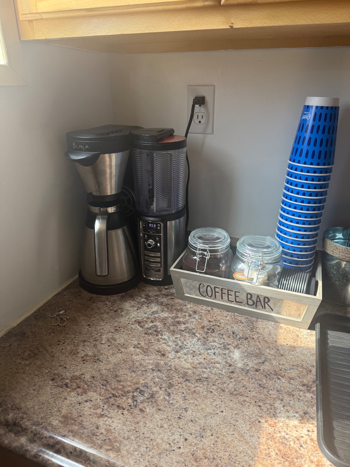 A corner of the kitchen showcases a coffee maker next to a blender on a granite countertop. Various glass jars are neatly arranged beside a decorative container labeled 'COFFEE BAR,' accompanied by a stack of colorful cups.