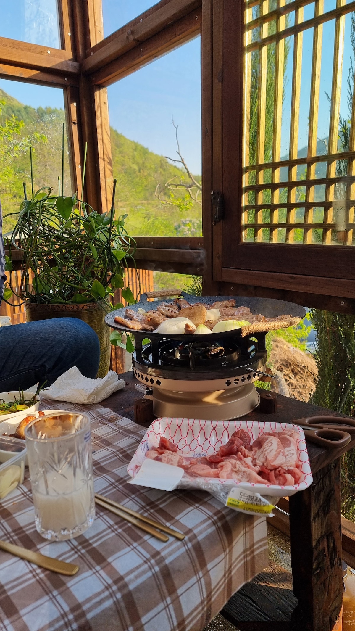 A cozy sunroom dining area features a portable grill placed on a small table, surrounded by delicious food. Natural light filters through wooden window slats, illuminating greenery outside. A glass of beverage sits nearby, with a checkered tablecloth completing the inviting setting.