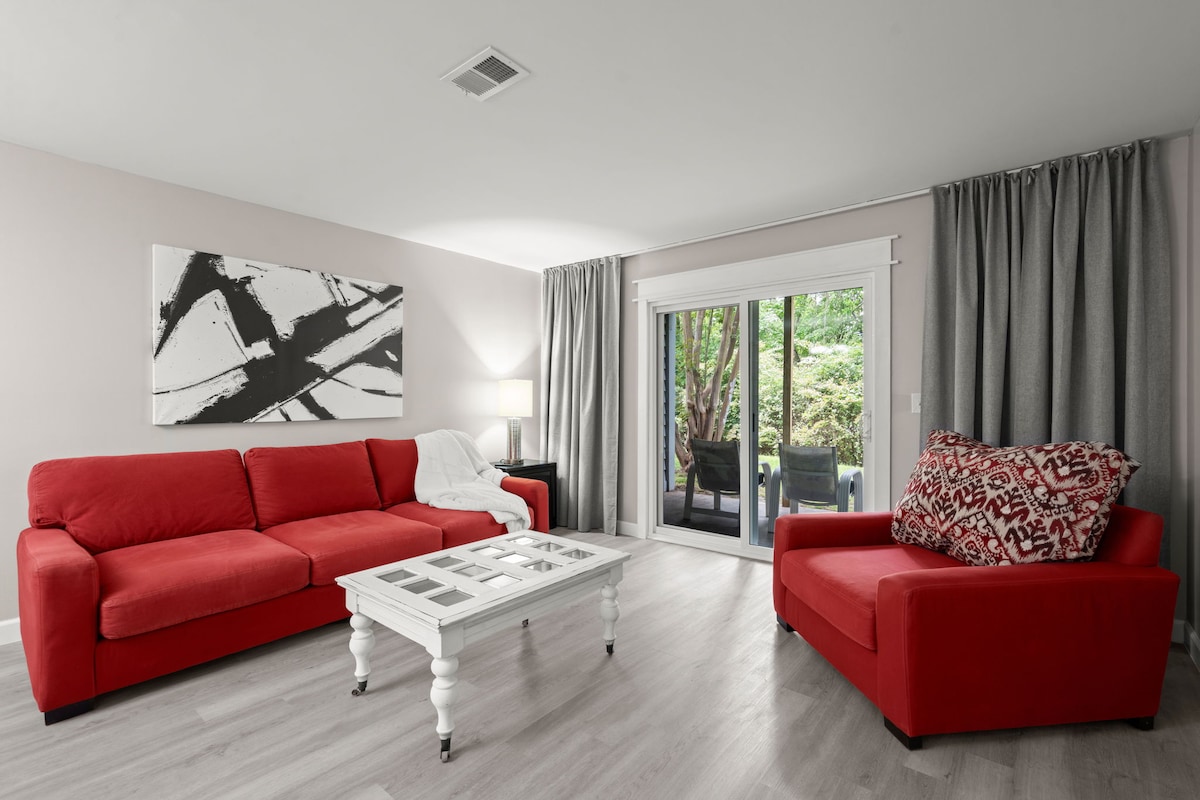 A lively living area features vibrant red seating, including a spacious sofa and an accent chair. A white coffee table sits in front, while large sliding glass doors lead to a private outdoor space surrounded by greenery. Soft curtains frame the windows, adding to the room's comfort.