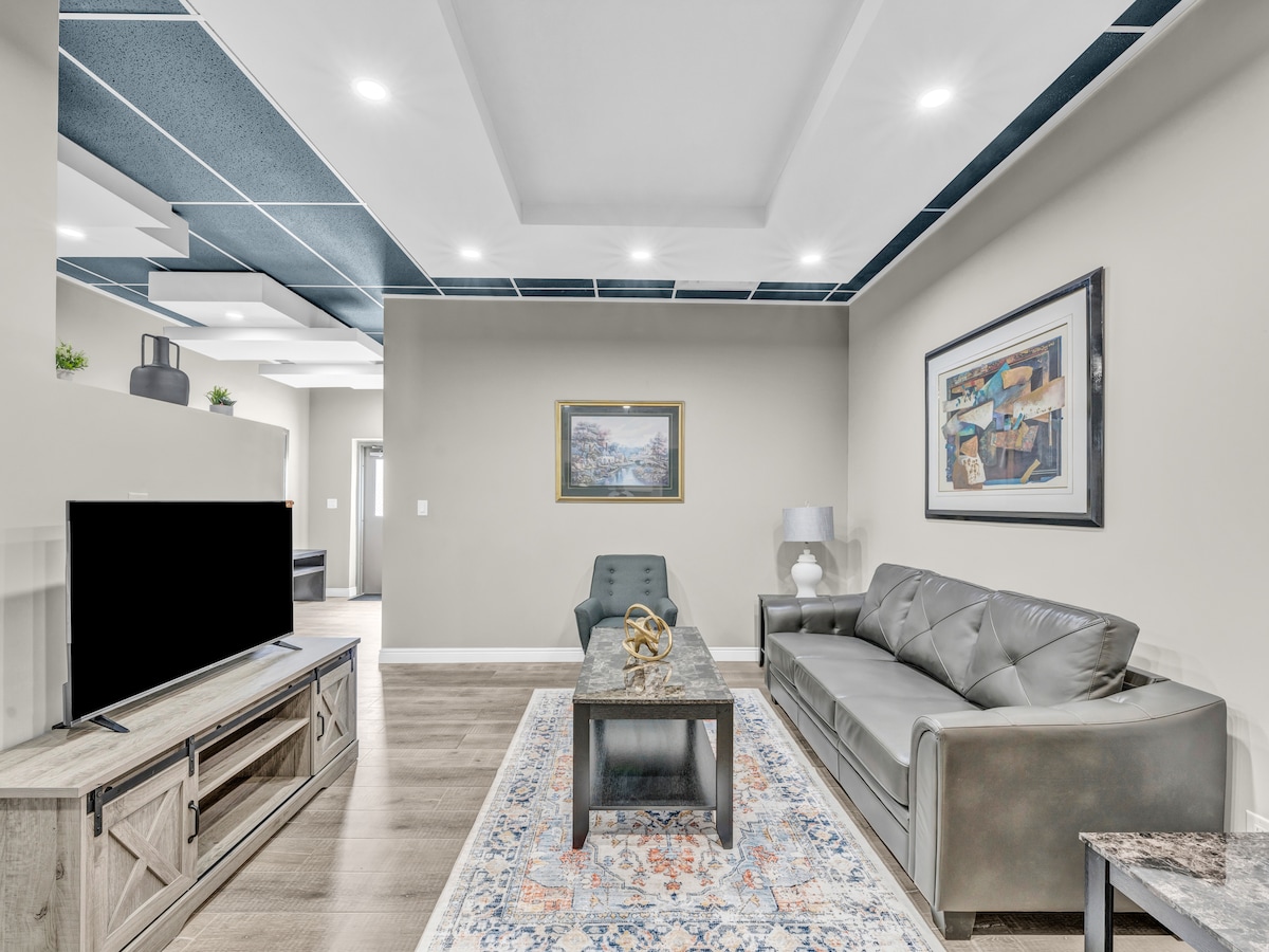 The living area features modern furnishings, including a sleek gray sofa and an accent chair. A large Smart TV is positioned on a stylish media unit. Natural light fills the space from recessed lighting and nearby openings, highlighting a decorative area rug and minimalist decor.