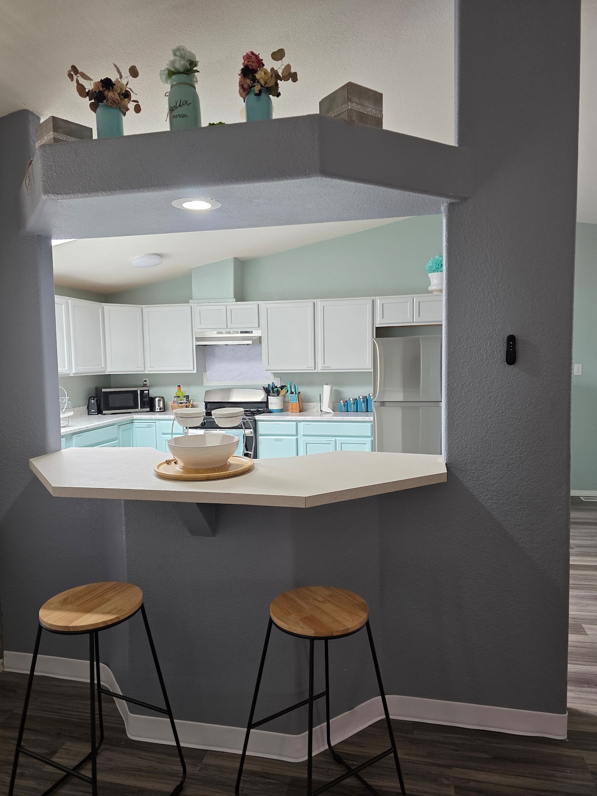 A kitchen area features a bar counter with two wooden stools. Light blue cabinets complement a white countertop, with kitchen appliances visible in the background. Decorative items are placed on the bar, adding a touch of color to the space.
