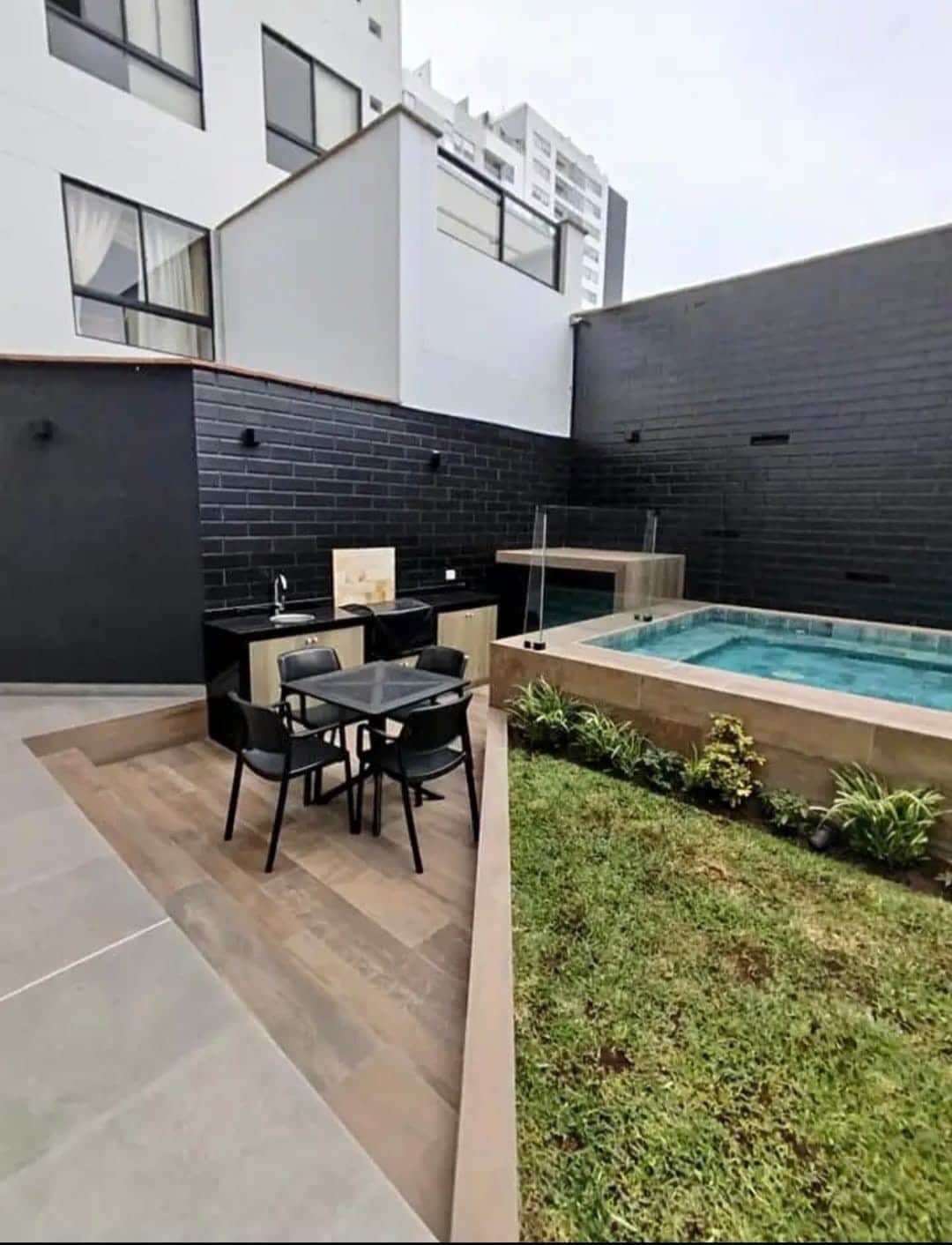 An outdoor area features a small dining table with four black chairs beside a modern swimming pool. The surrounding space includes a lawn and neatly arranged plants, while the walls exhibit a sleek dark finish, contributing to the contemporary design.