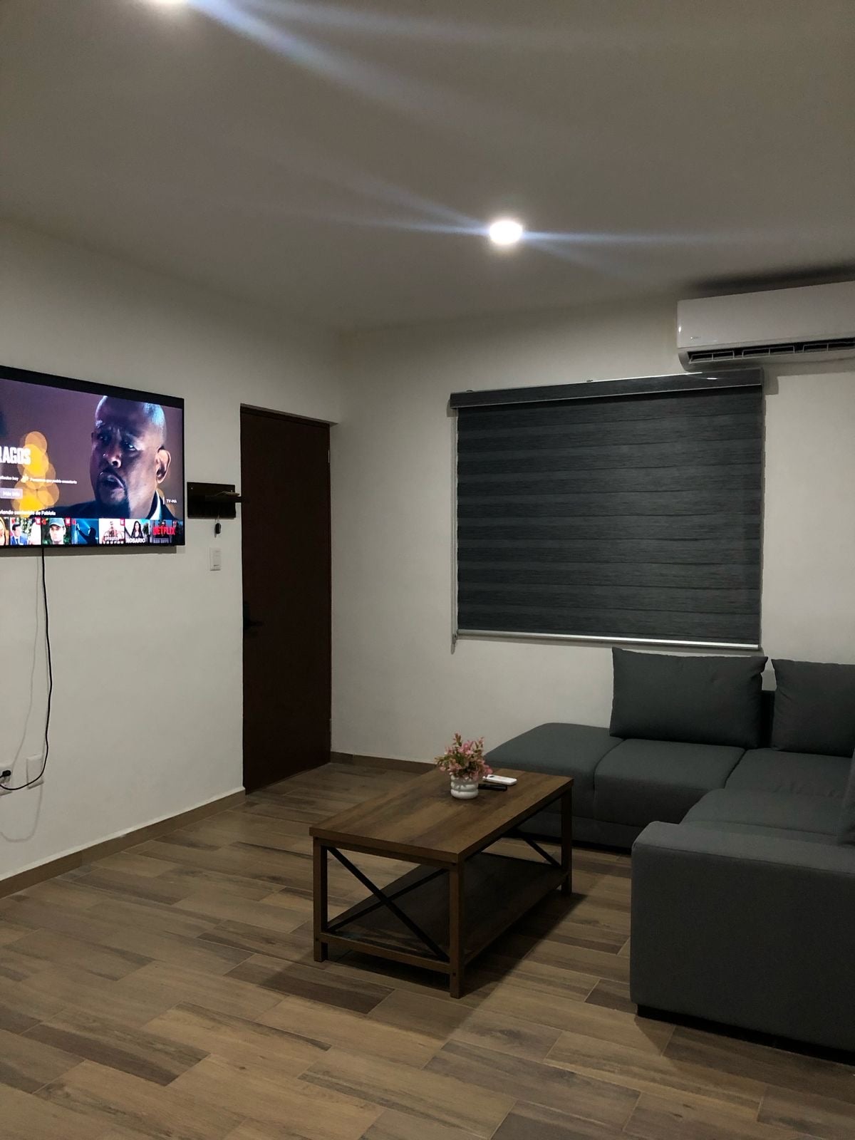 A modern living area features a dark grey sectional sofa positioned near a wooden coffee table. A flat-screen television is mounted on the wall, while a window with a dark roller shade allows for controlled lighting. The floor is covered with light-toned wooden planks, enhancing the space.