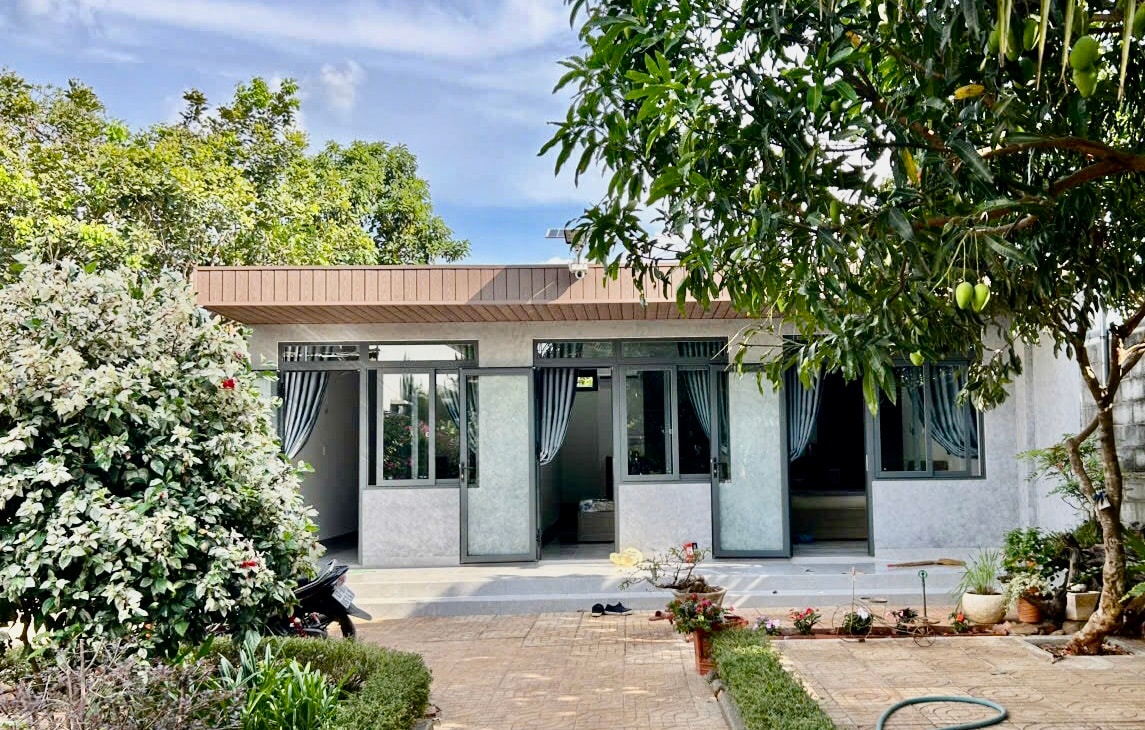 The exterior of the property is presented, showcasing a single-story structure with large glass windows framed by sheer curtains. Surrounding greenery includes shrubs and trees, adding a natural element to the setting. A paved pathway leads to the entrance, enhancing accessibility.