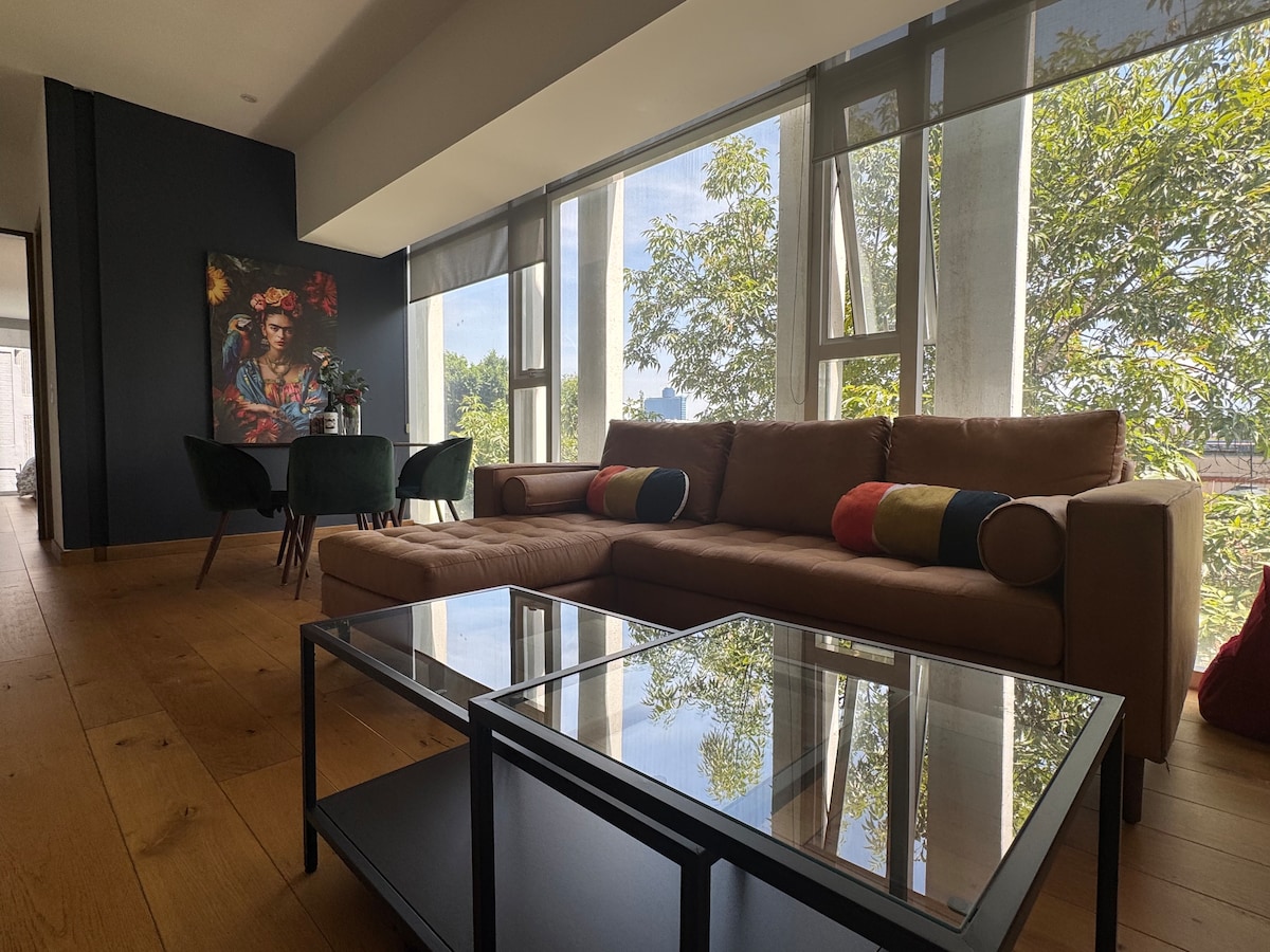 A spacious living area is adorned with a modern sofa, featuring colorful cushions. A sleek glass coffee table sits in front of the sofa, reflecting natural light from large windows framed by sheer curtains. A dining table and chairs are visible in the background.