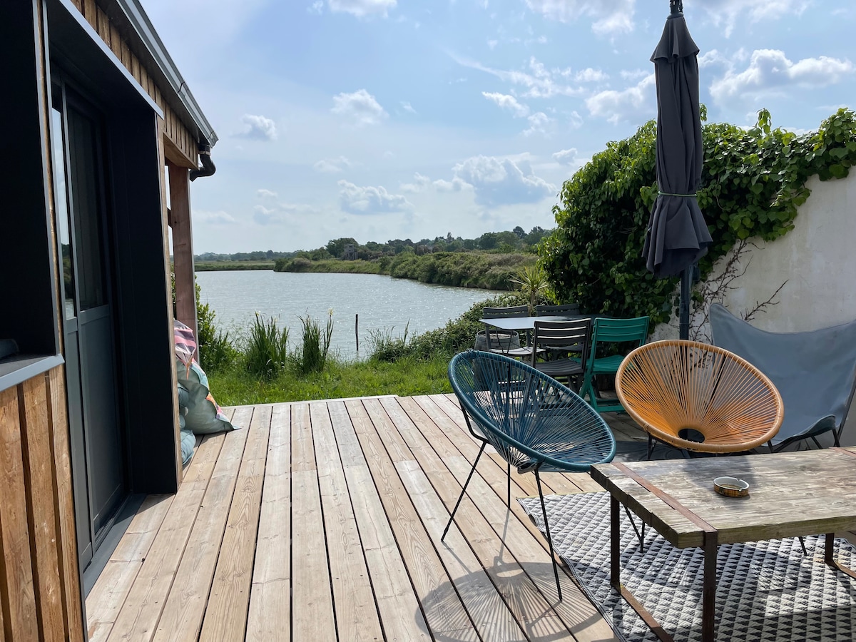 A spacious terrace is presented, featuring a weathered wooden deck that overlooks a peaceful marsh area. Two colorful lounge chairs and a table are arranged, with greenery surrounding the space. A large umbrella provides shade, complementing the serene atmosphere next to the water.