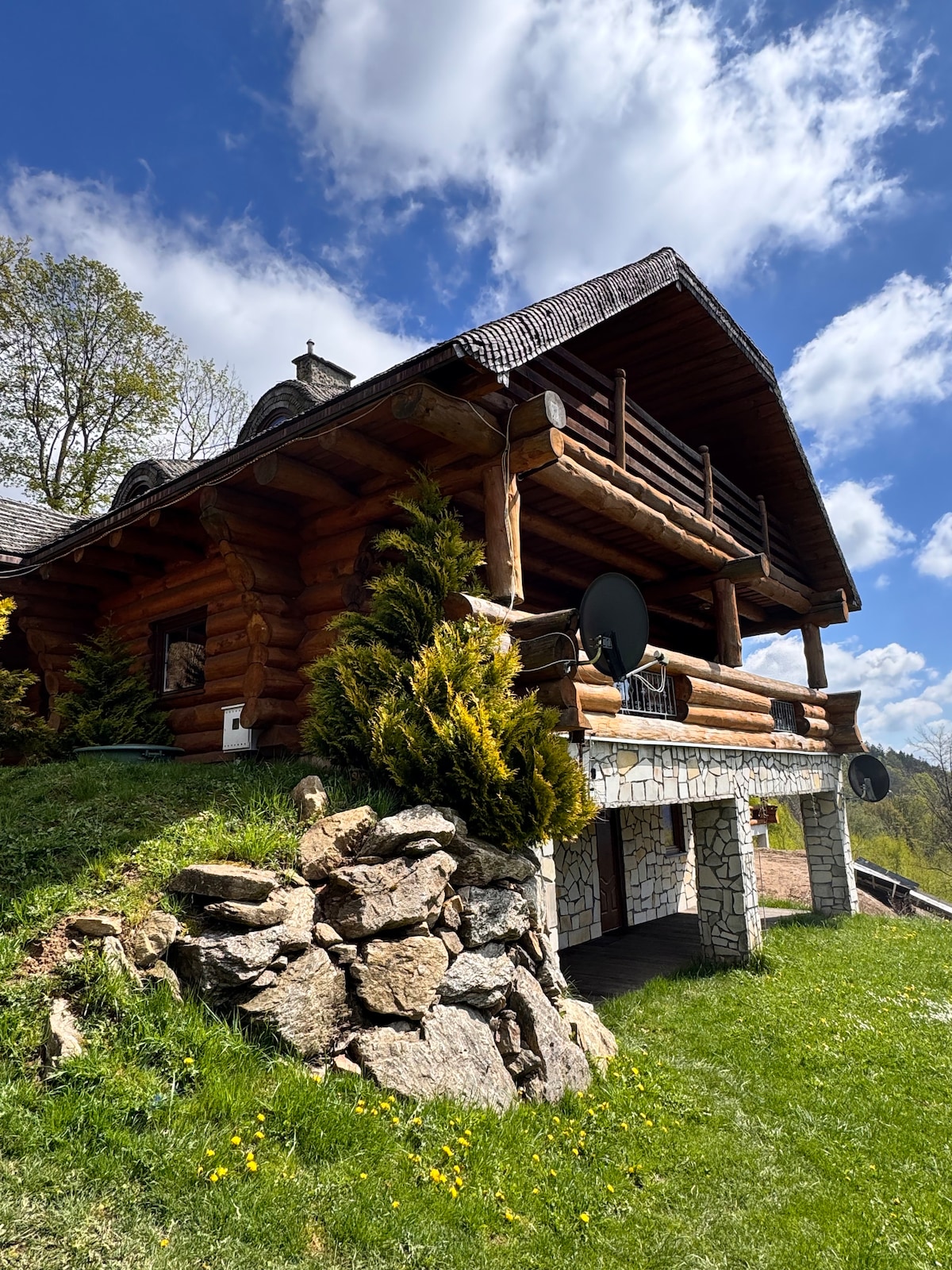 A rustic wooden cabin with a sloped roof is surrounded by a lush green hillside. Smooth rocks define parts of the landscape, and a well-maintained lawn features small yellow flowers. The terrace showcases wooden railings and is equipped with satellite dishes, complementing the natural setting.