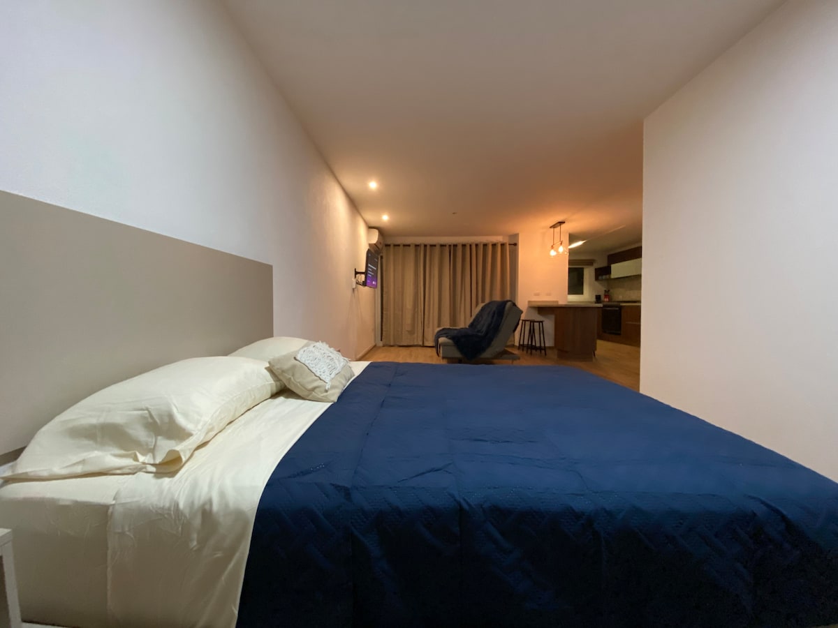 A spacious bedroom features a comfortable bed with crisp linens and a navy blue coverlet. The room is brightened by warm lighting, and a cozy seating area with a reclining chair is visible in the background. Sliding glass doors open to a natural light-filled space.