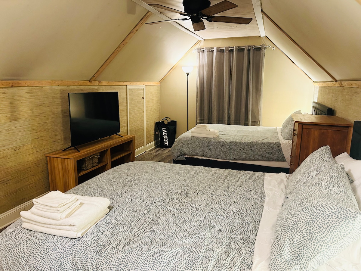 A spacious sleeping area is presented featuring two beds with light grey patterned bedding. A flat-screen TV is mounted on the wall opposite the beds. Natural light enters through a window covered by sheer curtains, and a floor lamp provides additional illumination.