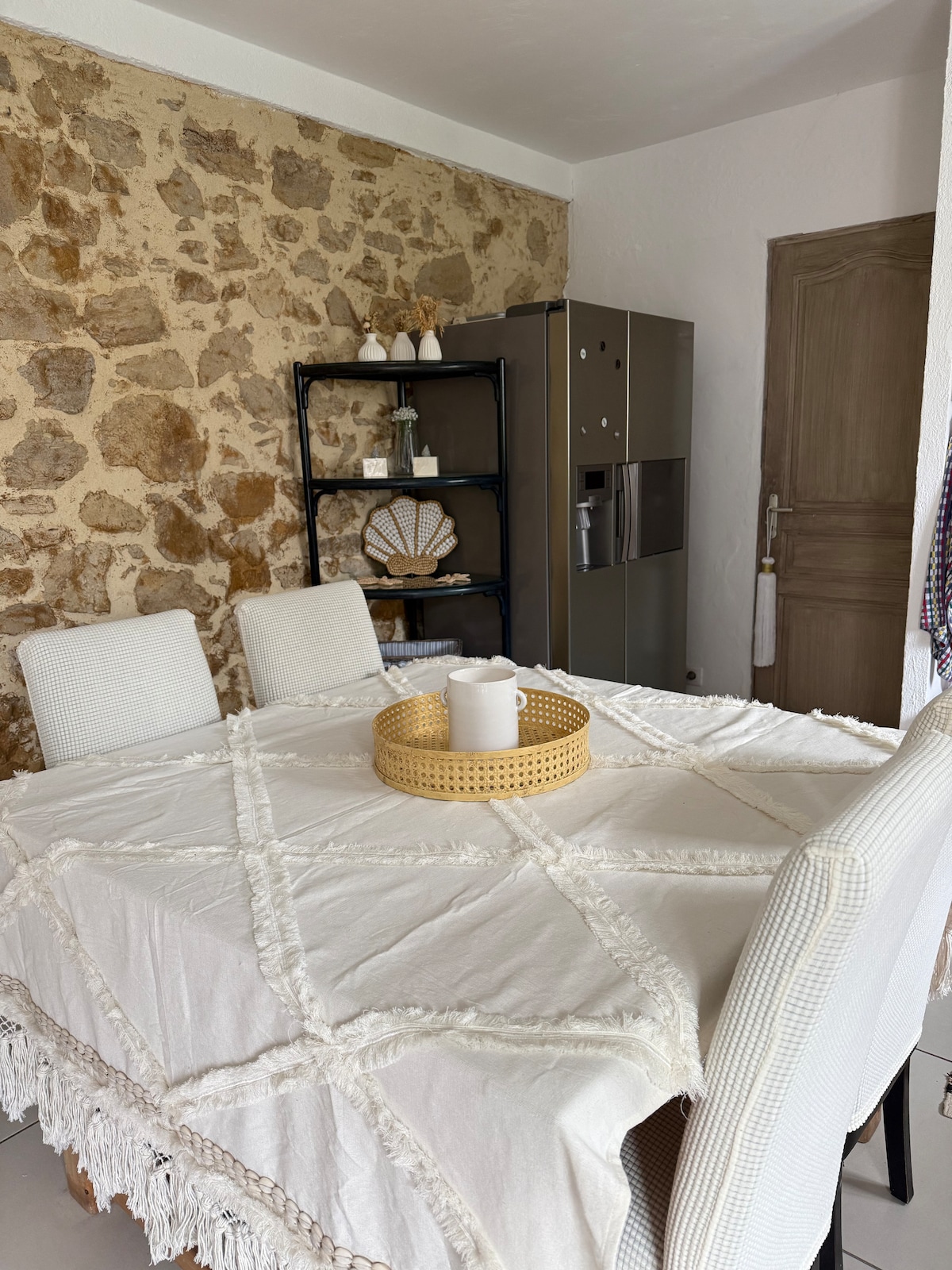 A dining area is presented with a textured stone wall. A round, woven tray sits at the center of a rectangular table, draped with a fringed tablecloth. Four upholstered chairs surround the table, while a shelving unit displays decorative items against the adjacent wall.
