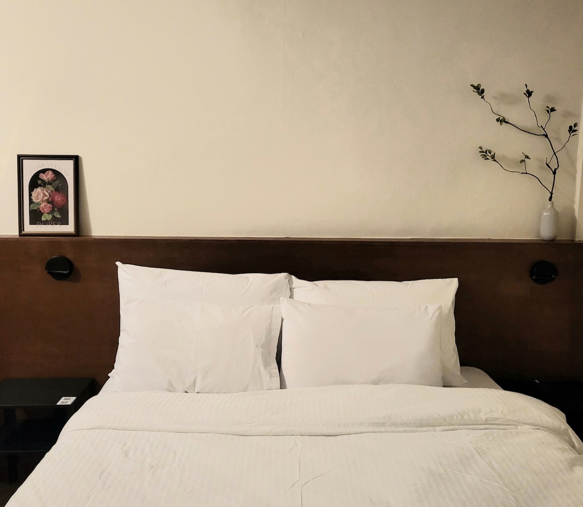 A serene sleeping area is presented, featuring a spacious bed adorned with crisp white linens. Beside the bed, a small nightstand holds a decorative frame with floral art. A minimalist branch arrangement in a vase adds a touch of nature to the wall.