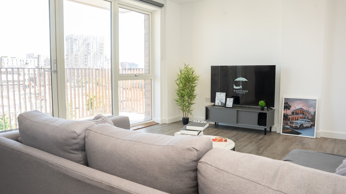 A bright living area features a comfortable gray sofa arranged to face a modern TV on a low entertainment unit. A small plant adds a touch of greenery, while natural light streams in through large sliding doors leading to a private balcony.
