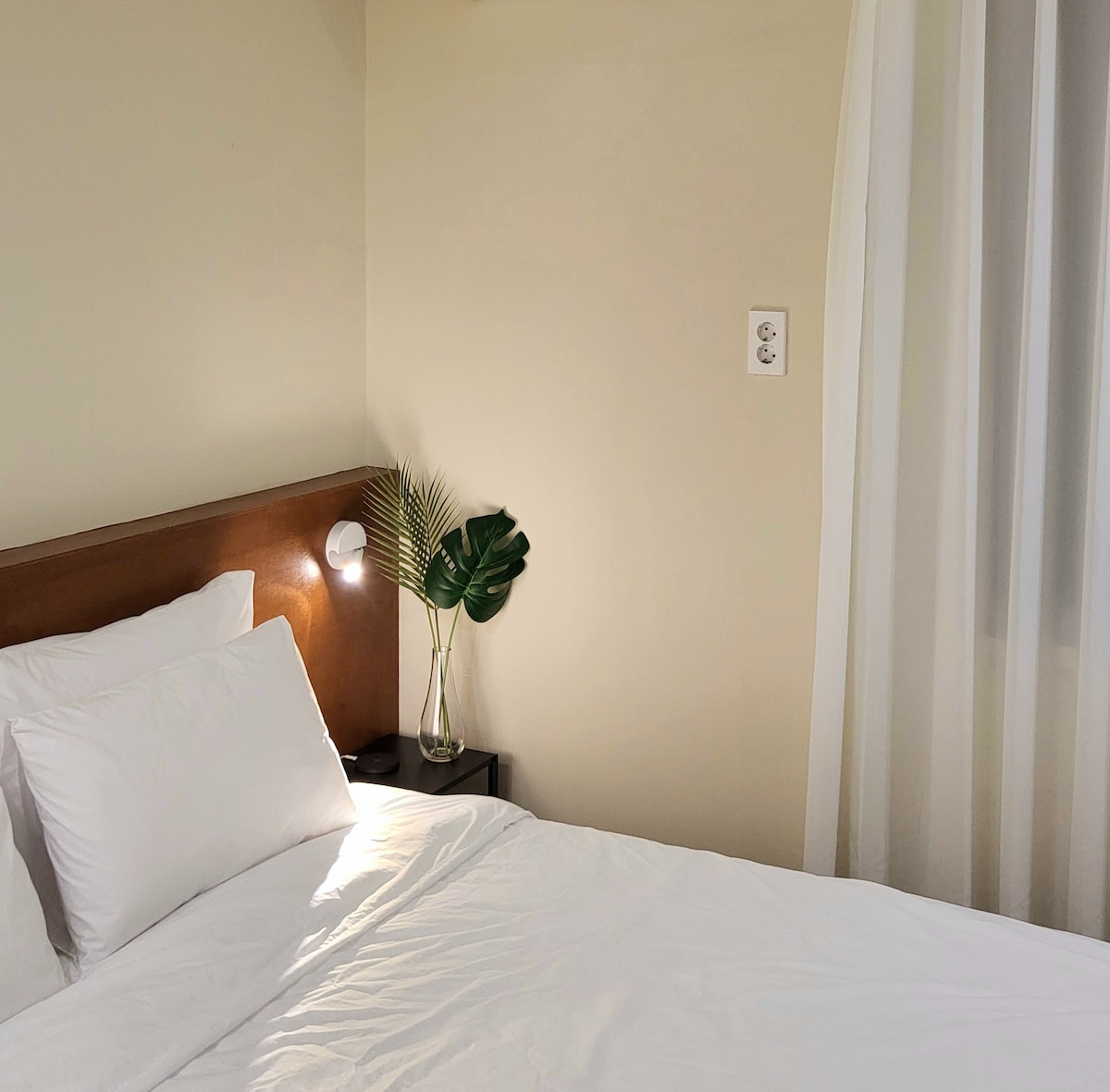 A serene sleeping area is presented with a spacious bed dressed in white linens. A bedside table hosts a stylish lamp and a small vase containing a green plant. Soft curtains diffuse natural light, enhancing the calming ambiance.