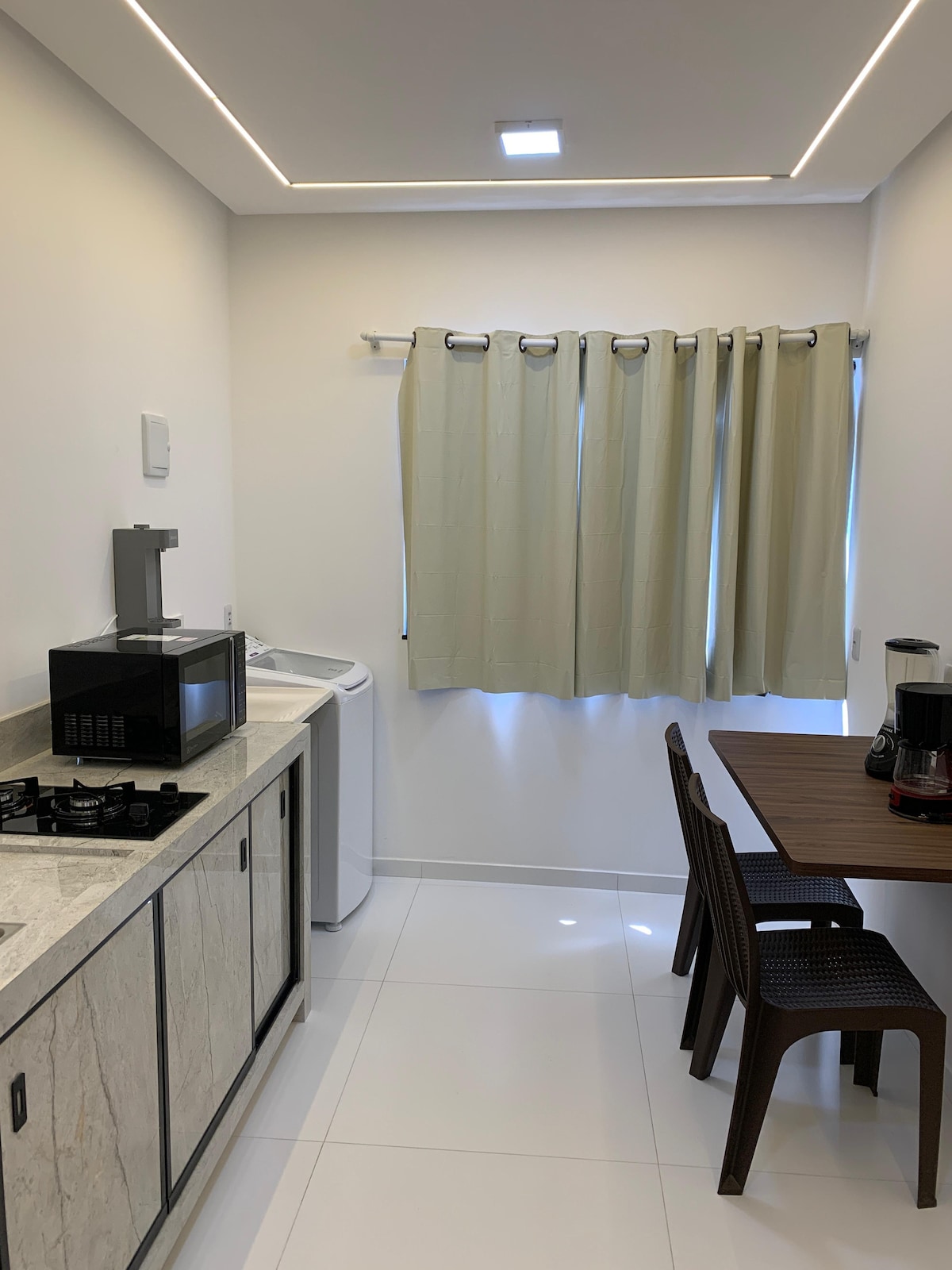 A compact kitchen area features a marble countertop with a gas stove, microwave, and coffee maker. A small wooden table accompanies two chairs, while a washing machine is positioned beside a window covered by light-colored curtains, allowing natural light to brighten the space.