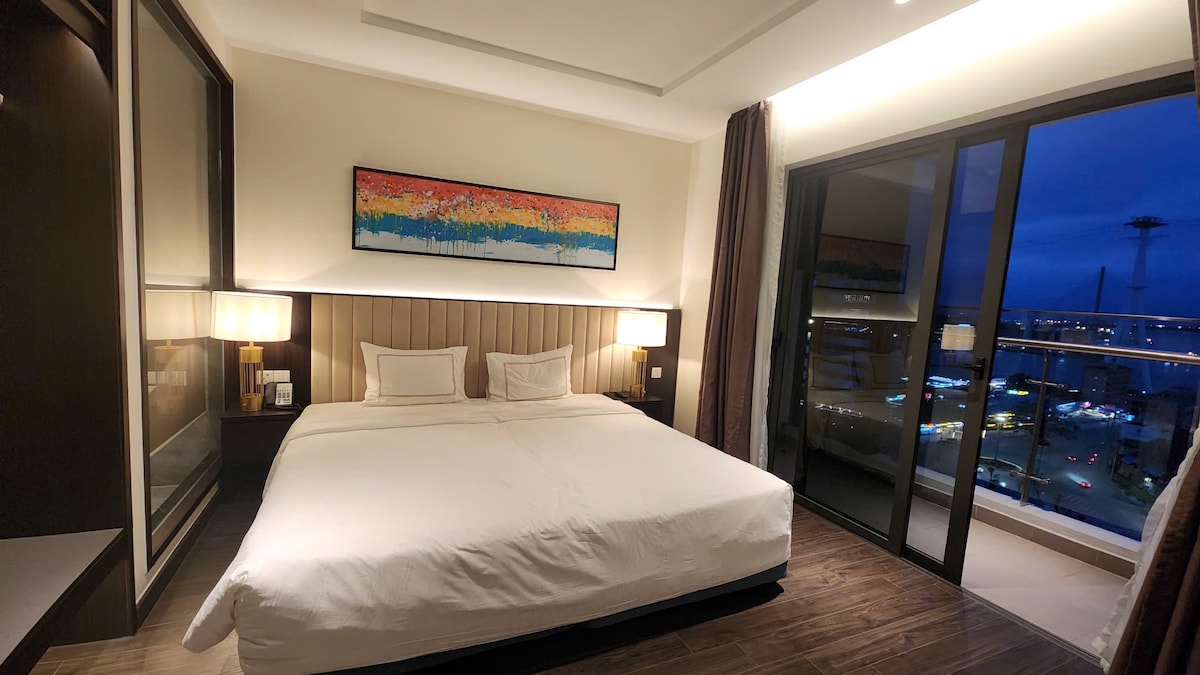 A spacious sleeping area features a large bed with crisp white linens, framed by two bedside lamps. A colorful piece of wall art adds a touch of character. Floor-to-ceiling windows reveal a city view, enhancing the room's modern design.