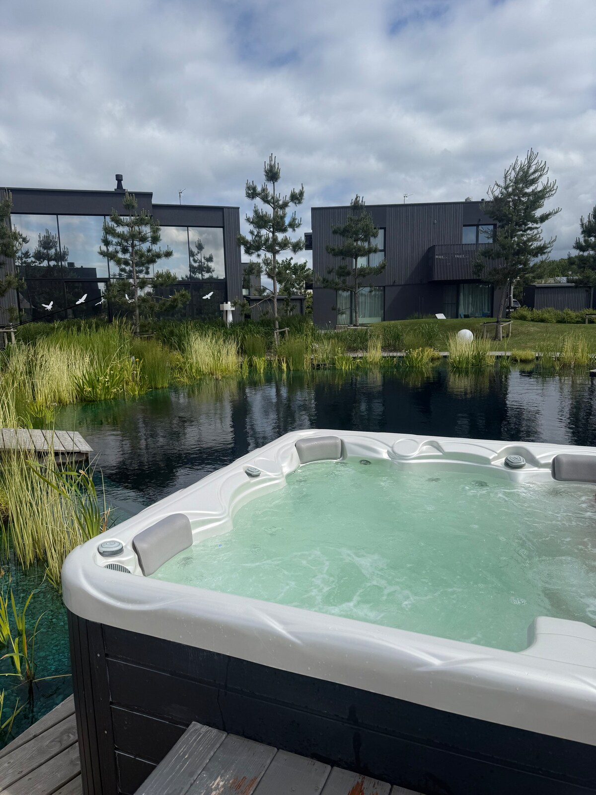 A hot tub is positioned on a wooden deck, surrounded by lush greenery and water features. The serene landscape includes modern multi-story buildings in the background, partially obscured by ornamental trees. The cloudy sky provides soft lighting, creating a calm atmosphere.